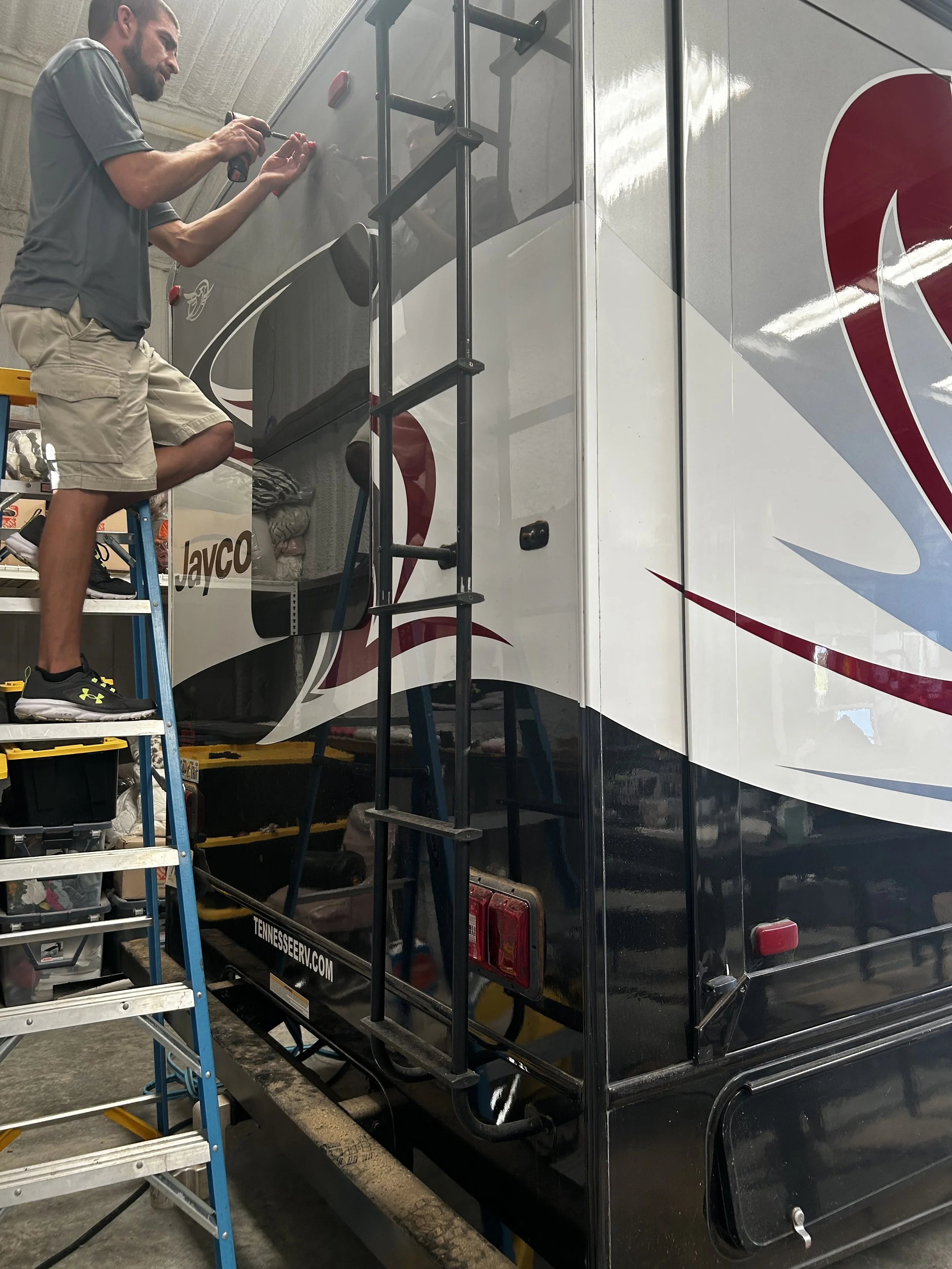 A man on a ladder working on the exterior of a large black and white recreational vehicle, possibly performing repairs or maintenance.