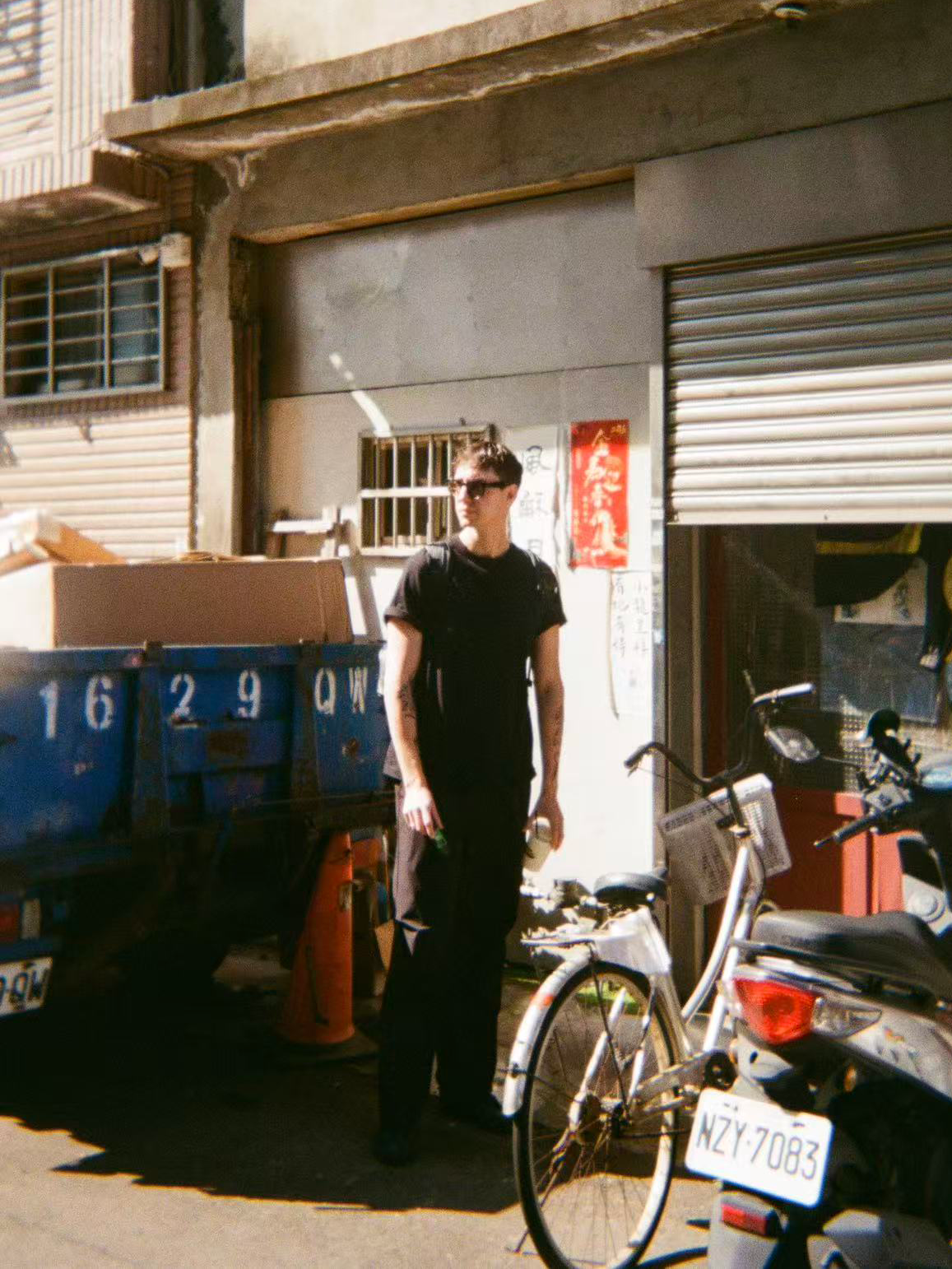 A man stands next to bicycles and a blue truck with a large cardboard box on it on a street in an urban area with storefronts and signage.