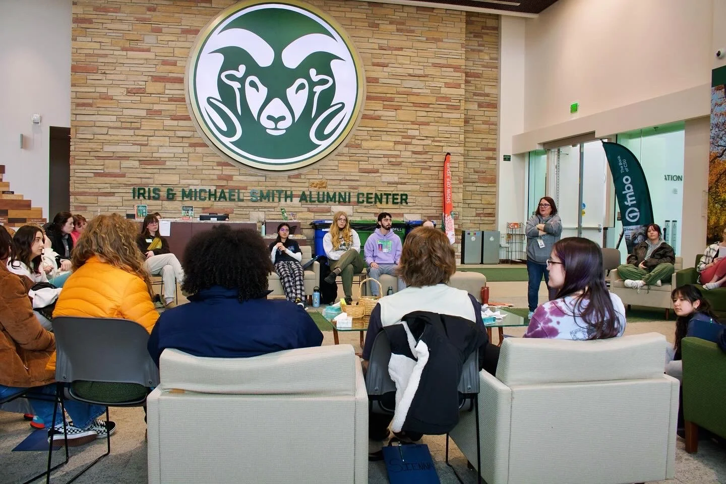 "Voices United"
A group of people sitting in a circle at Iris & Michael Smith Alumni Center, with a large green and white logo on the wall. The gathering appears to be a discussion or meeting.
