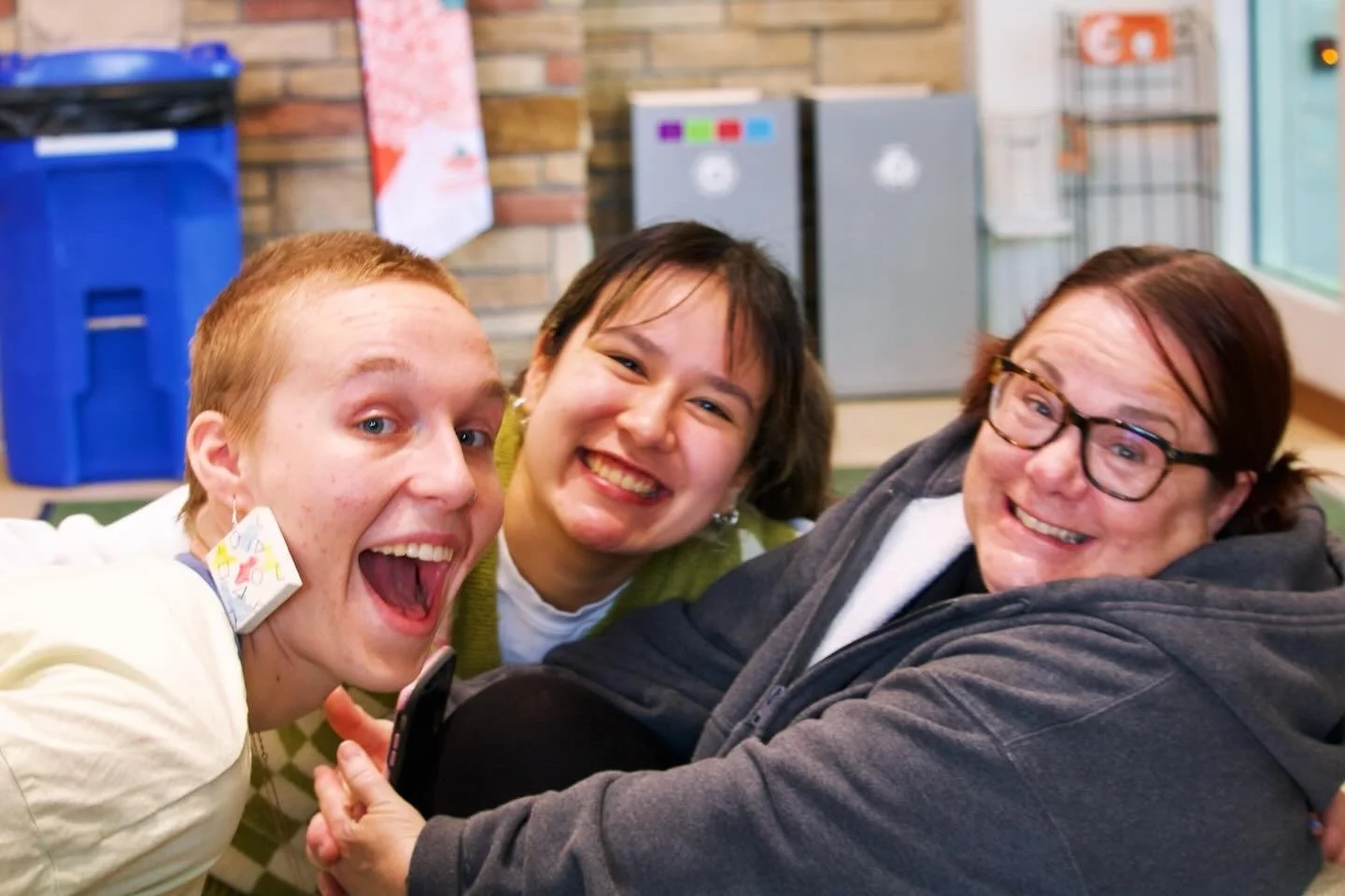 "Campus Friendships"
Three people smiling and posing together indoors, with a blue recycling bin and other objects in the background.