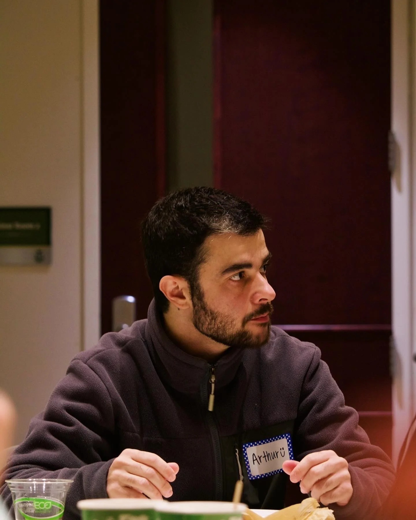 "Arthur"
Person sitting indoors wearing a dark jacket, looking to the side, with a name tag labeled 'Arthur' on their jacket.