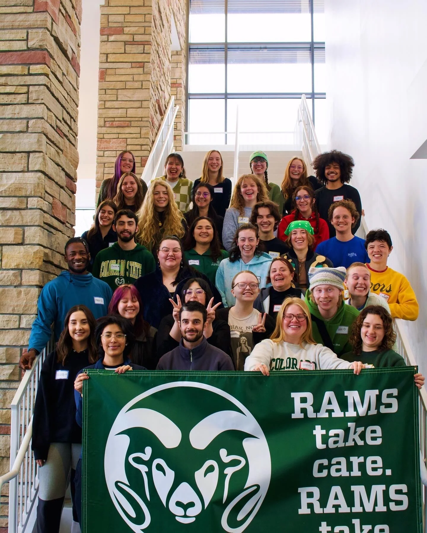 "Community First"
Group of people on stairs holding a green banner with a ram logo and the text 'Rams take care.' inside a building with stone walls.