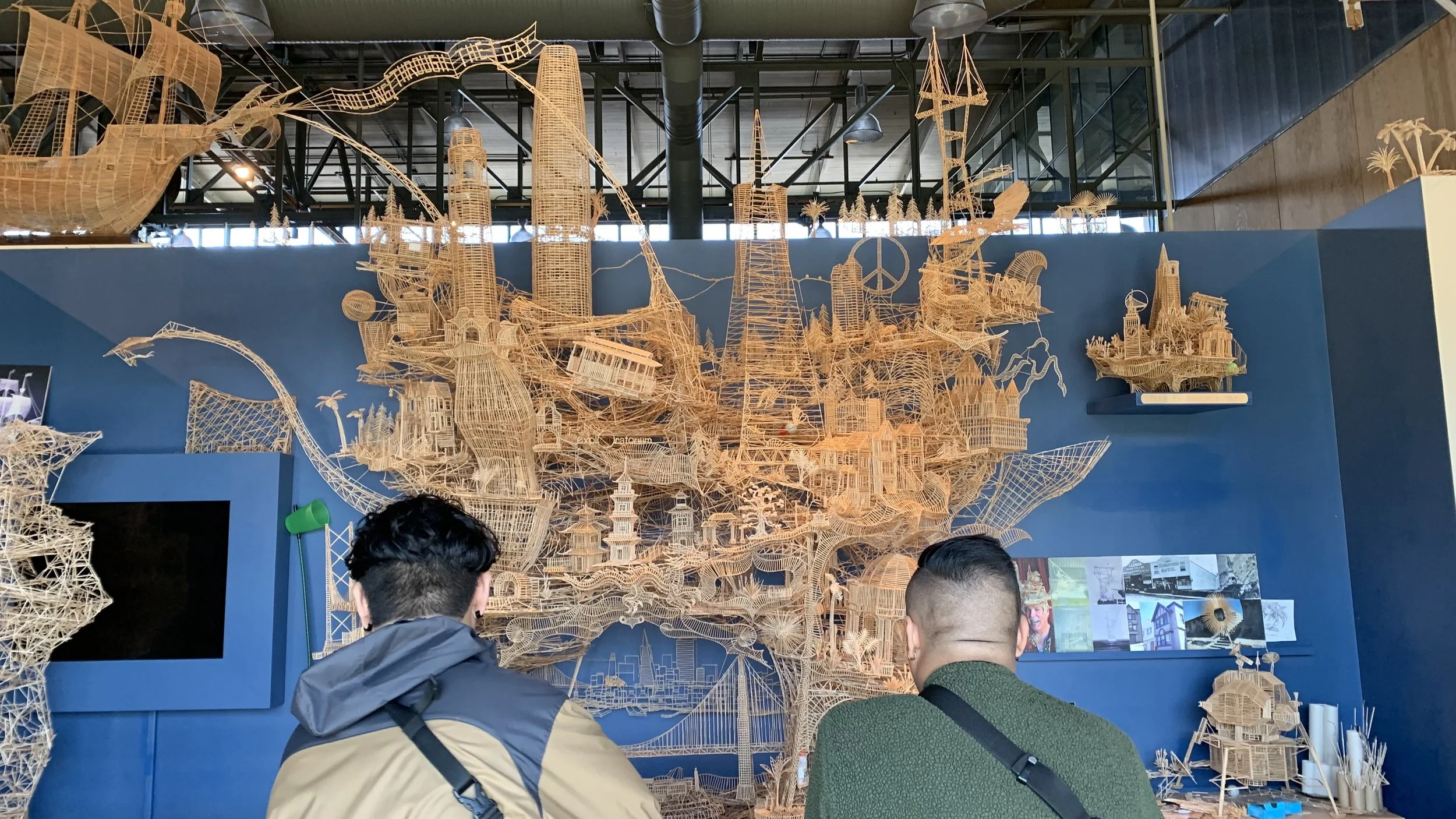 Two people viewing a large, intricate toothpick sculpture depicting famous landmarks, buildings, and scenes, displayed against a blue wall in a museum setting.