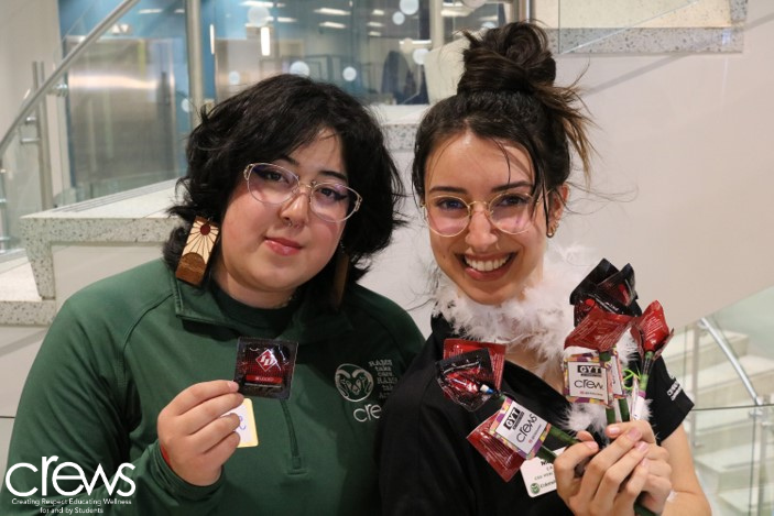 "Safe Choices"
Two people indoors holding and displaying a selection of condoms, standing in a modern, well-lit space with stairs in the background. They are wearing glasses and smiling towards the camera. The word 'crews' is visible in white text at