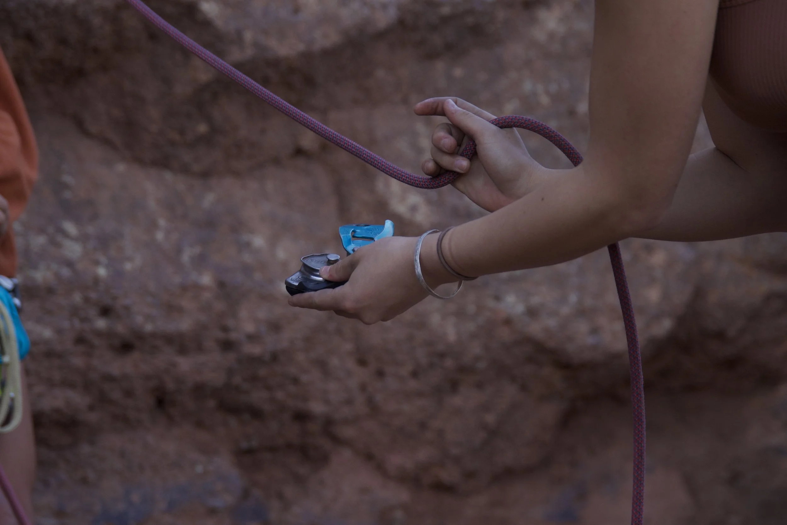 "Tools of the Trade"
Close-up of a person holding a climbing belay device and rope against a rock background.