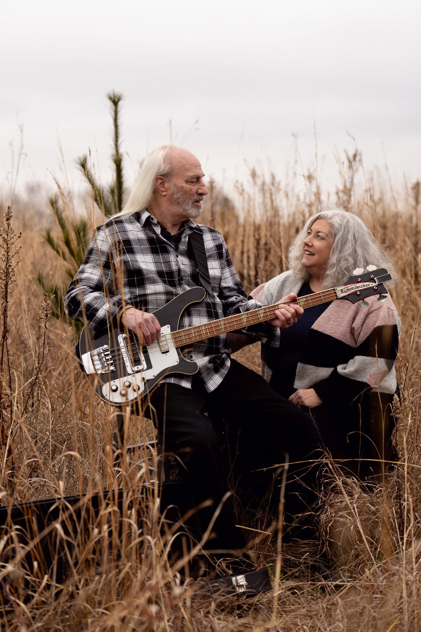 An elderly couple with long gray hair, sitting in a field of tall, dry grass. The man is playing an electric guitar, and the woman is smiling and looking at him, wearing a multicolored sweater.