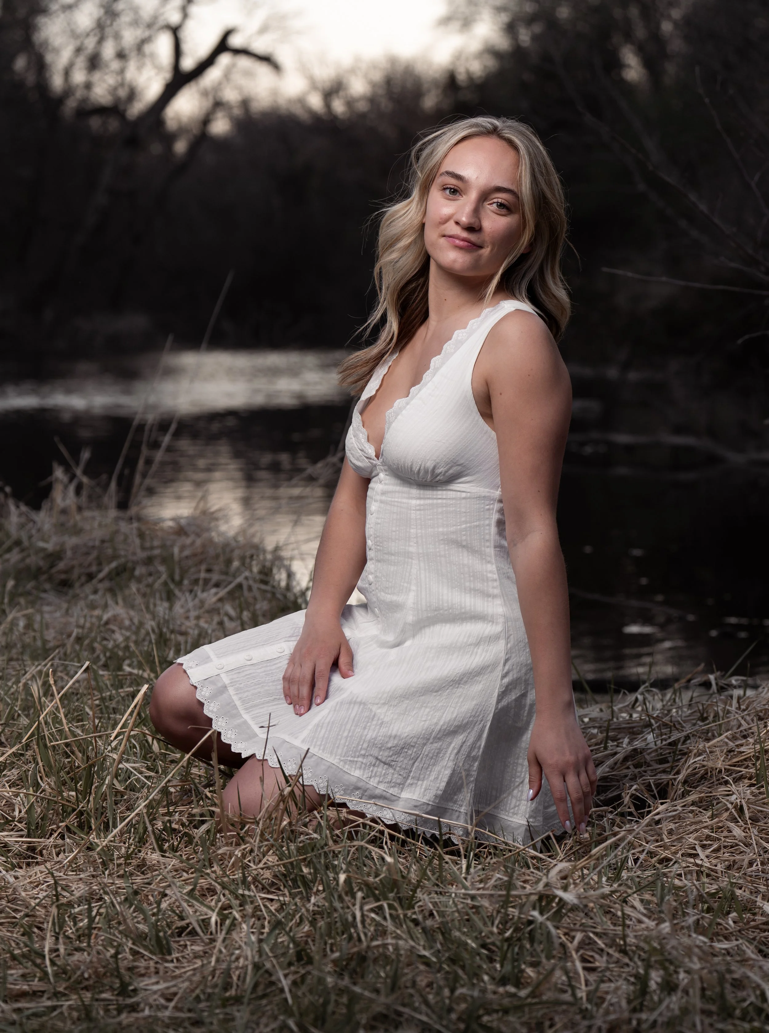 A young woman with wavy, shoulder-length blonde hair crouching outdoors in a field with dry grass and bare trees in the background, wearing a black long-sleeve shirt, a blue denim vest, a black skirt, and tan cowboy boots, smiling at the camera.