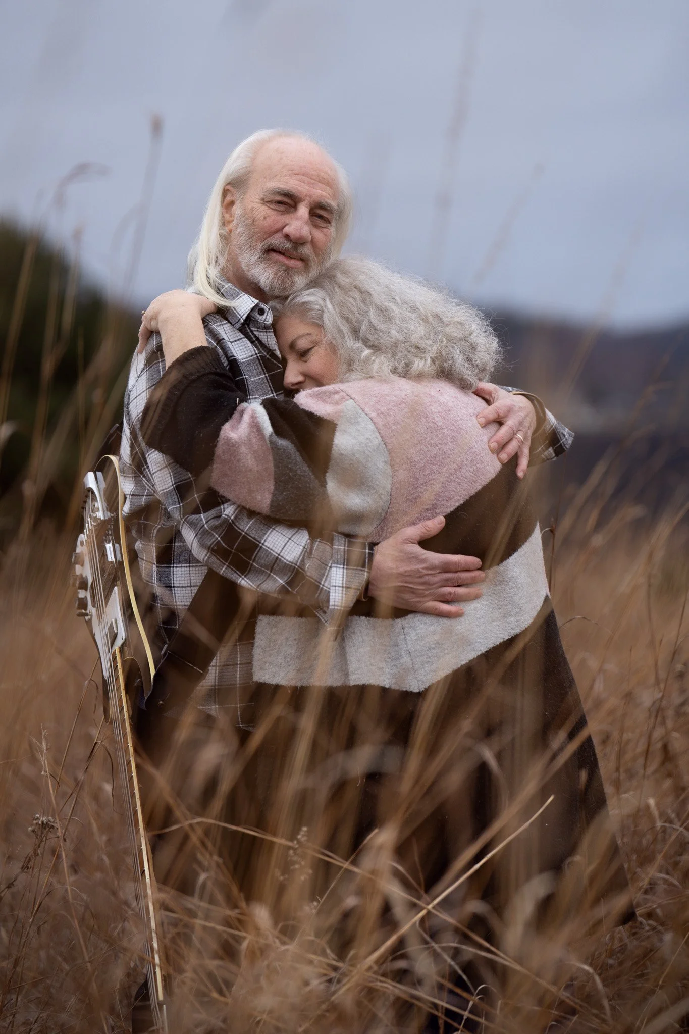 An elderly man and woman embrace in a field of tall grass, with the man holding a guitar, on a cloudy day.