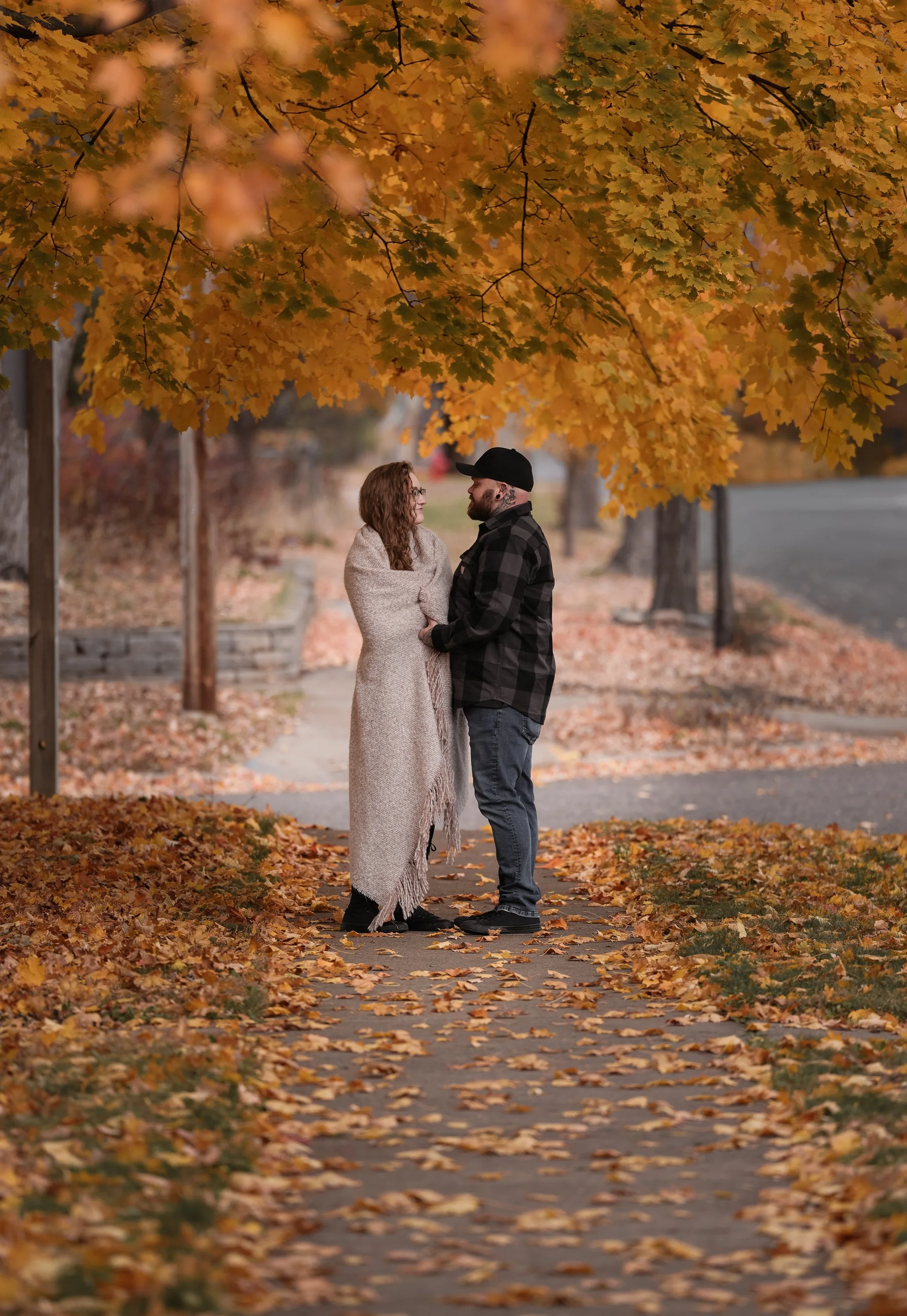 A couple standing on a leaf-covered sidewalk under a large tree with orange autumn leaves, gazing at each other.