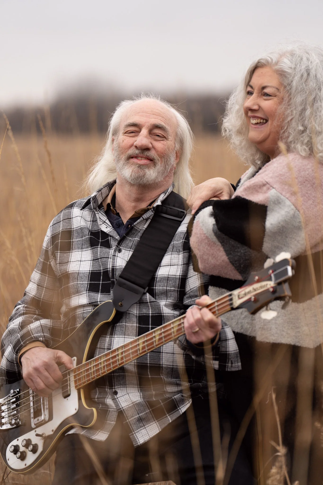 An elderly man and woman enjoying a moment together outdoors in a field. The man is holding an electric guitar and smiling, while the woman is smiling and touching his shoulder.