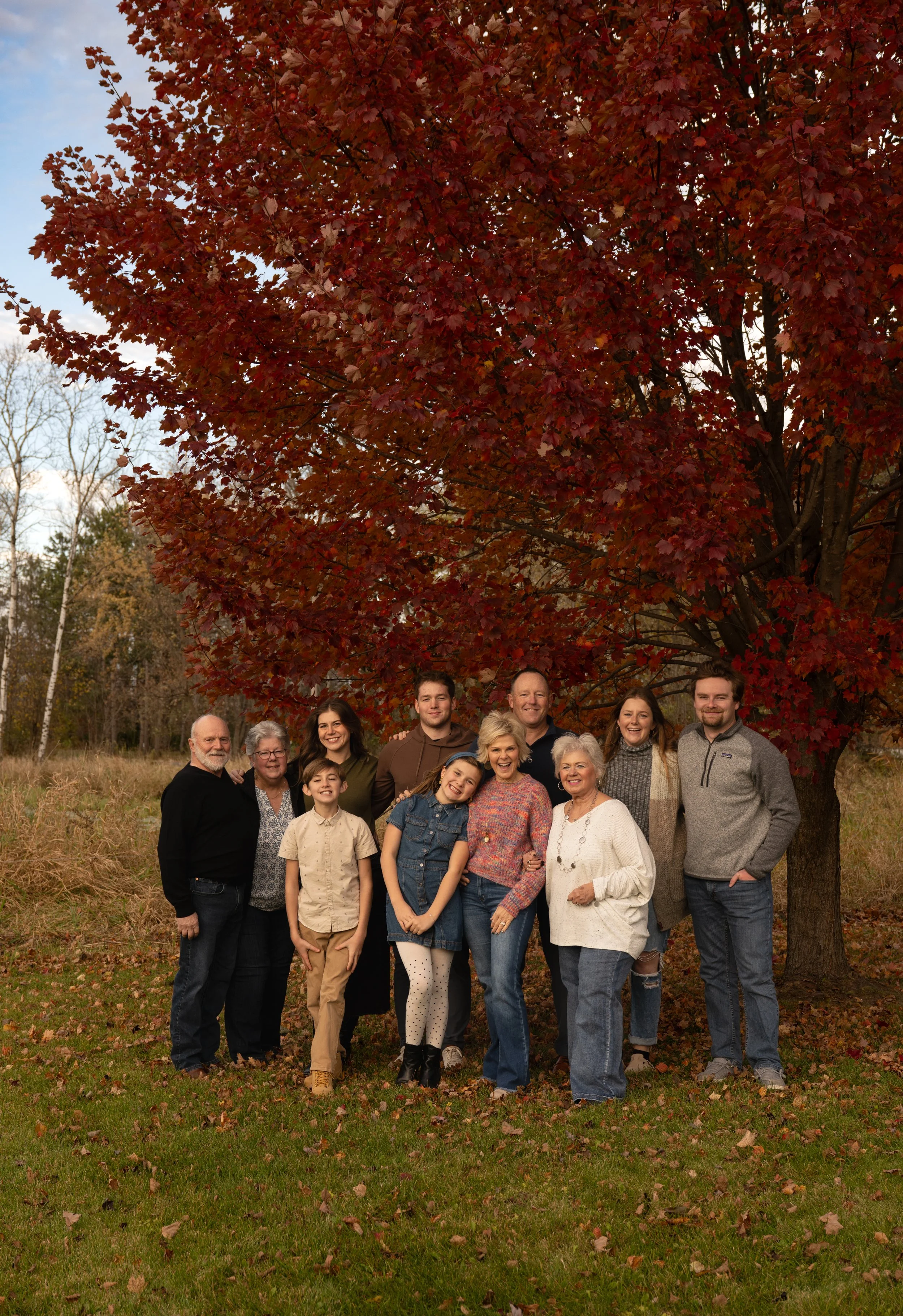 A multi-generational family portrait outdoors on a fall day, standing under a large red maple tree with fall foliage. The group includes children, adults, and elderly members smiling and embracing each other.