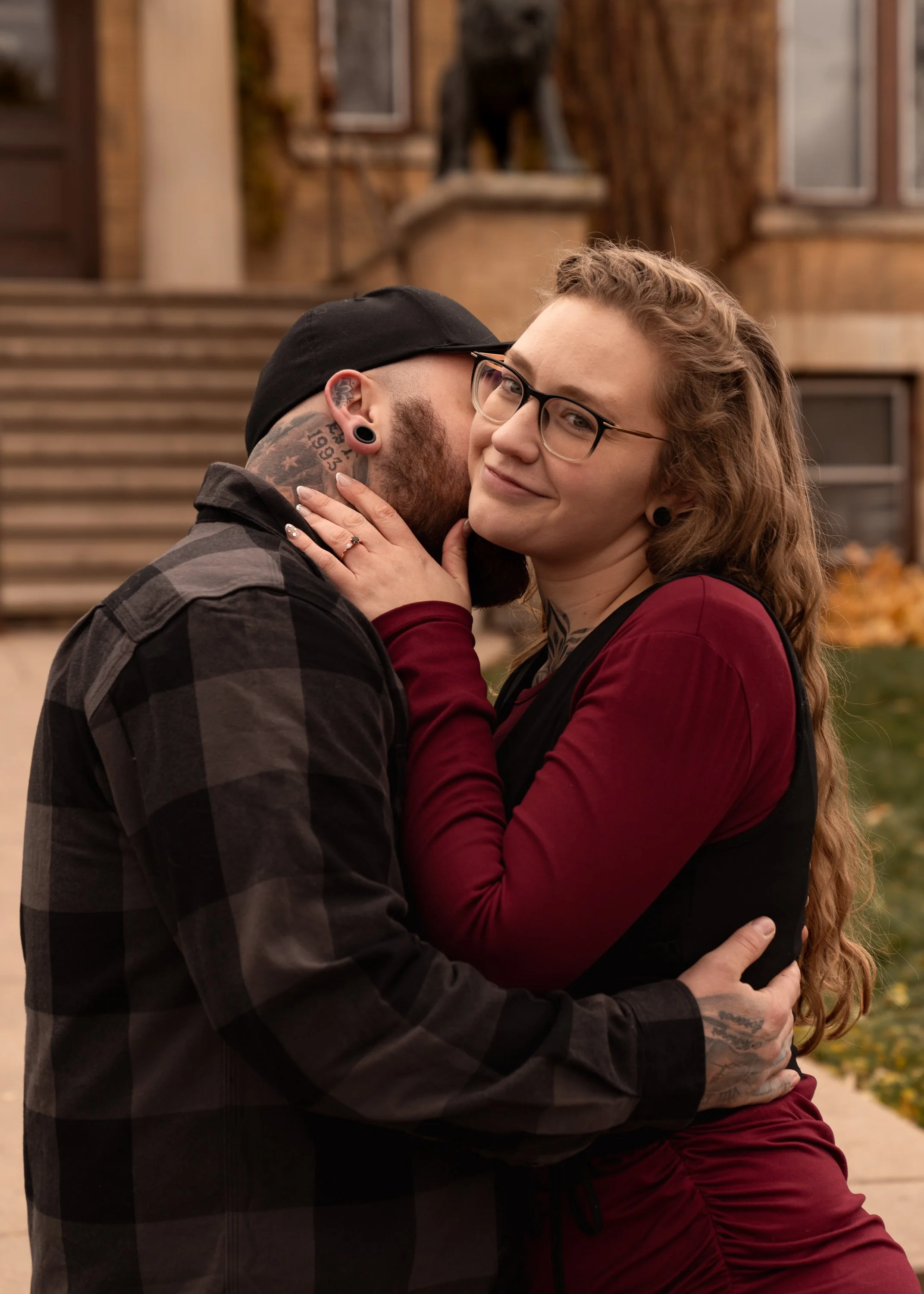 A couple hugging outdoors, with the woman smiling at the camera and the man whispering into her ear, in front of a house with stairs and autumn foliage.