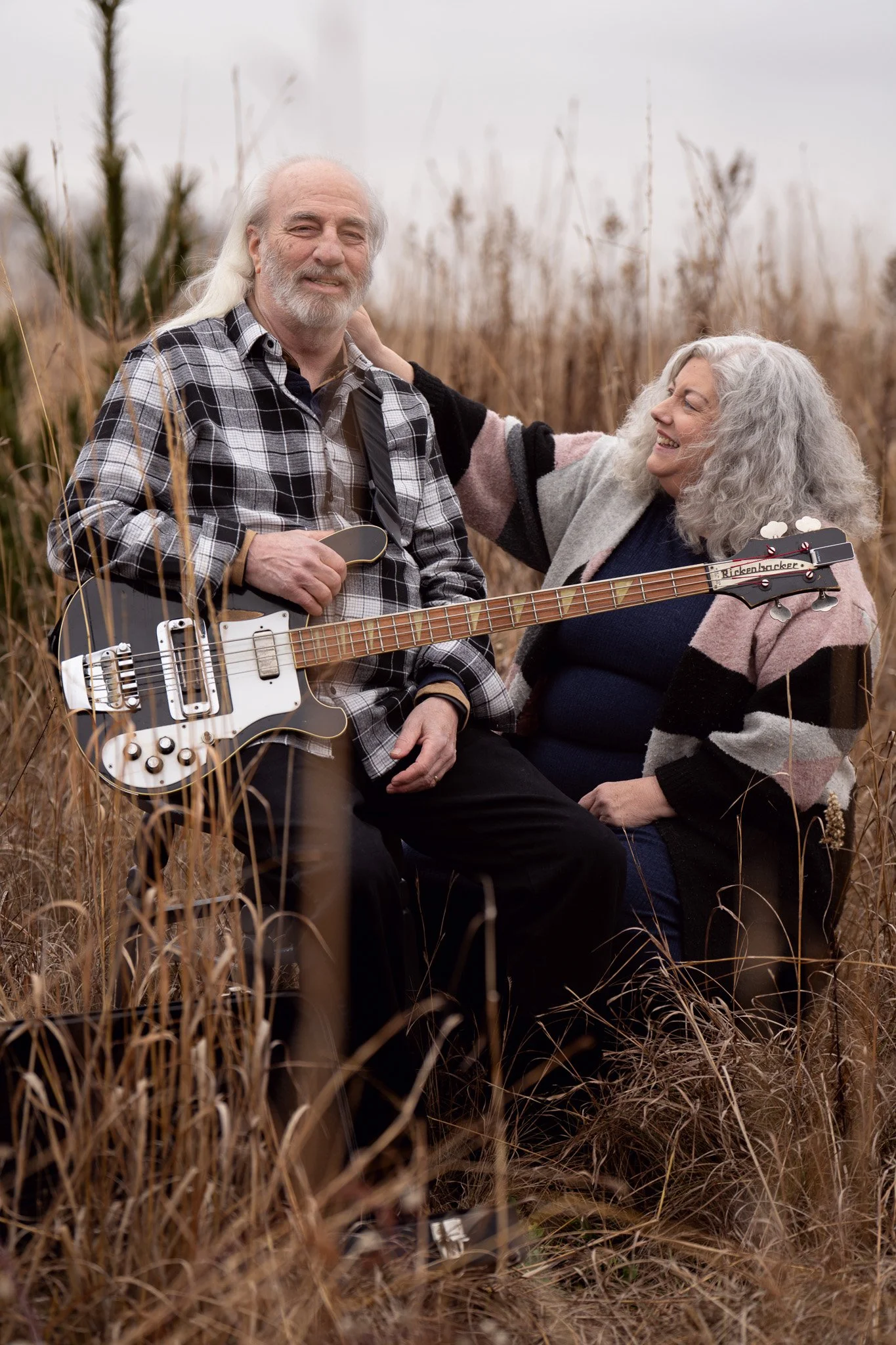 An elderly man and woman sitting in tall grass outdoors, with the man holding a bass guitar and the woman smiling and touching his shoulder.