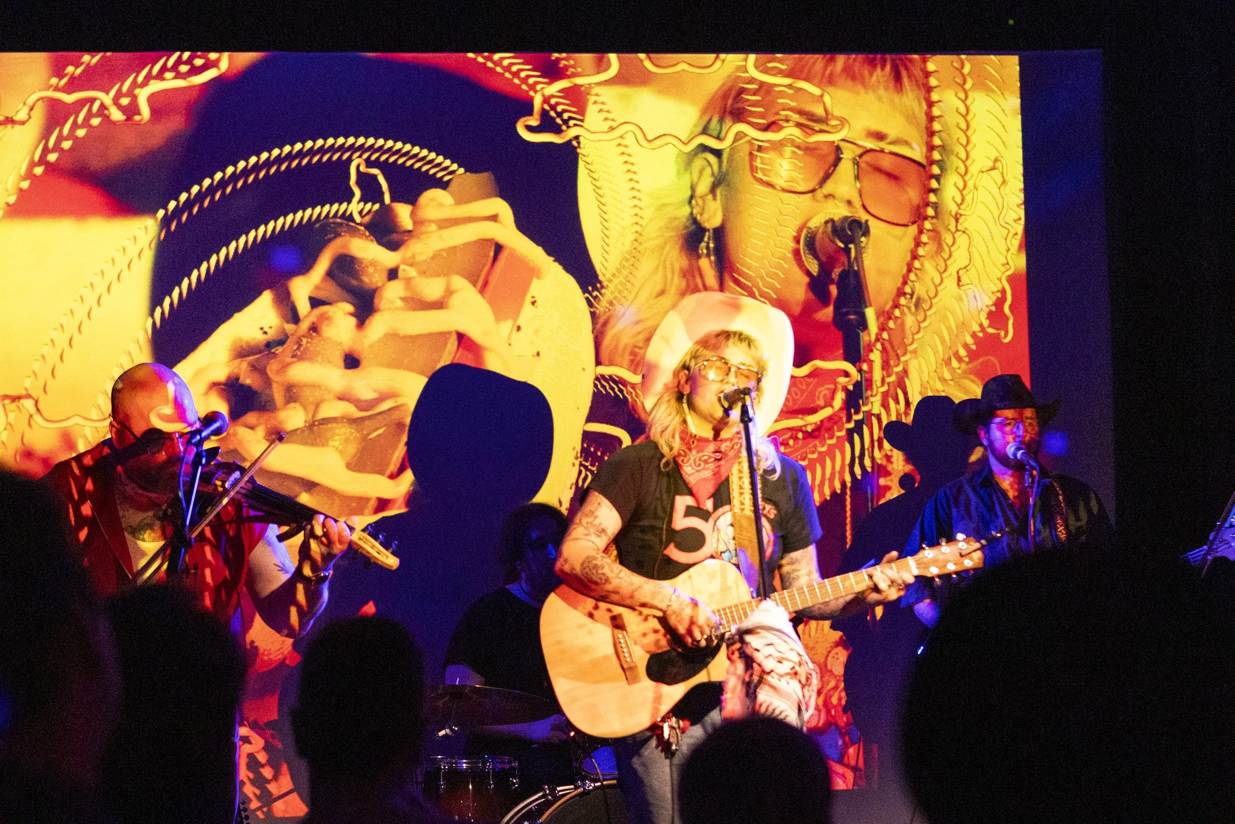 Musicians perform on stage with a bright, colorful background. The central figure is a woman with tattoos, wearing glasses, a cowboy hat, and a red bandana, playing an acoustic guitar and singing into a microphone. To her sides are other musicians, one playing a violin and another singing into a microphone with a cowboy hat.