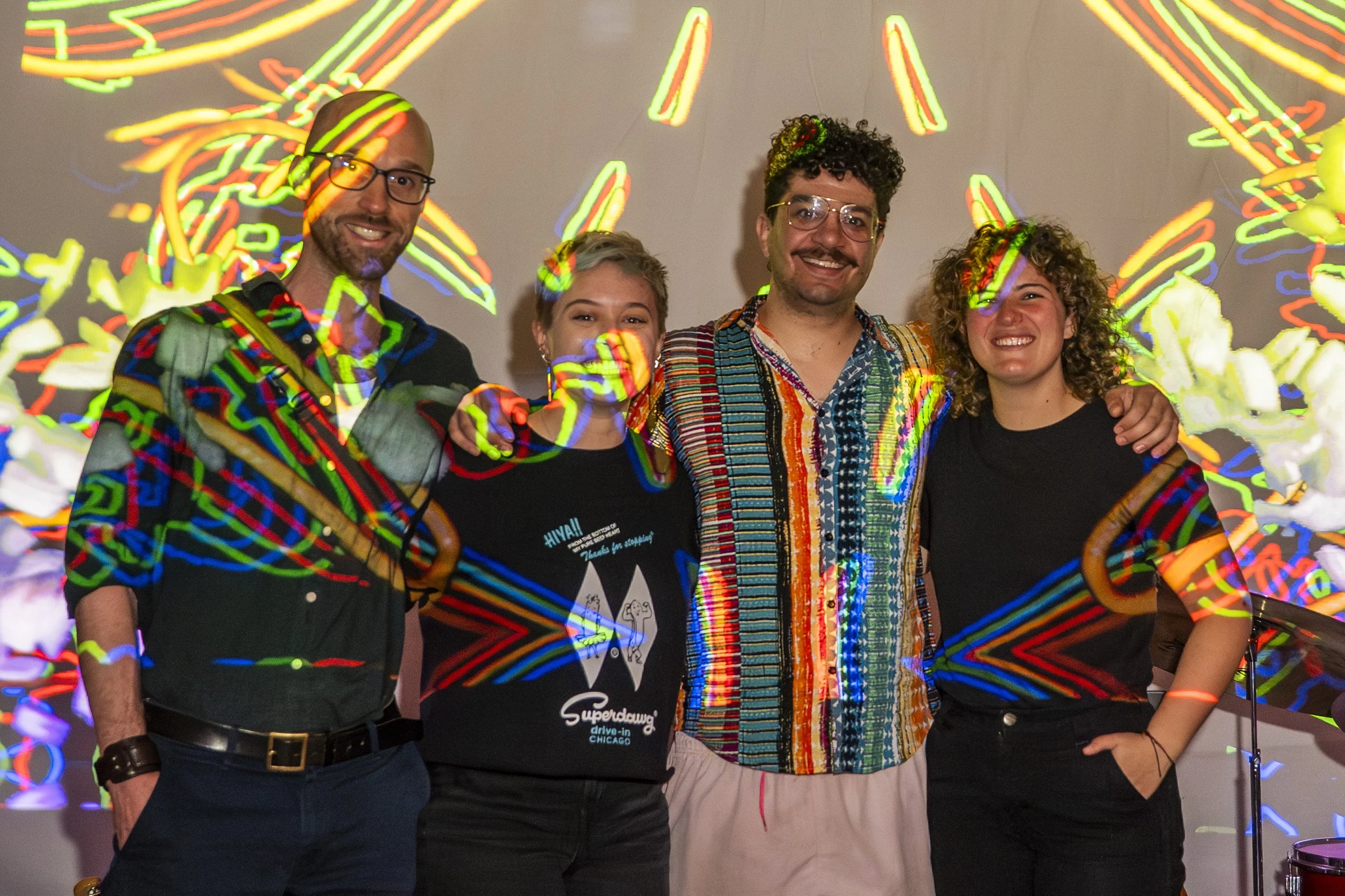Group of four people smiling and posing together at a colorful light art event with vibrant, multicolored light projections on the wall and their clothing.
