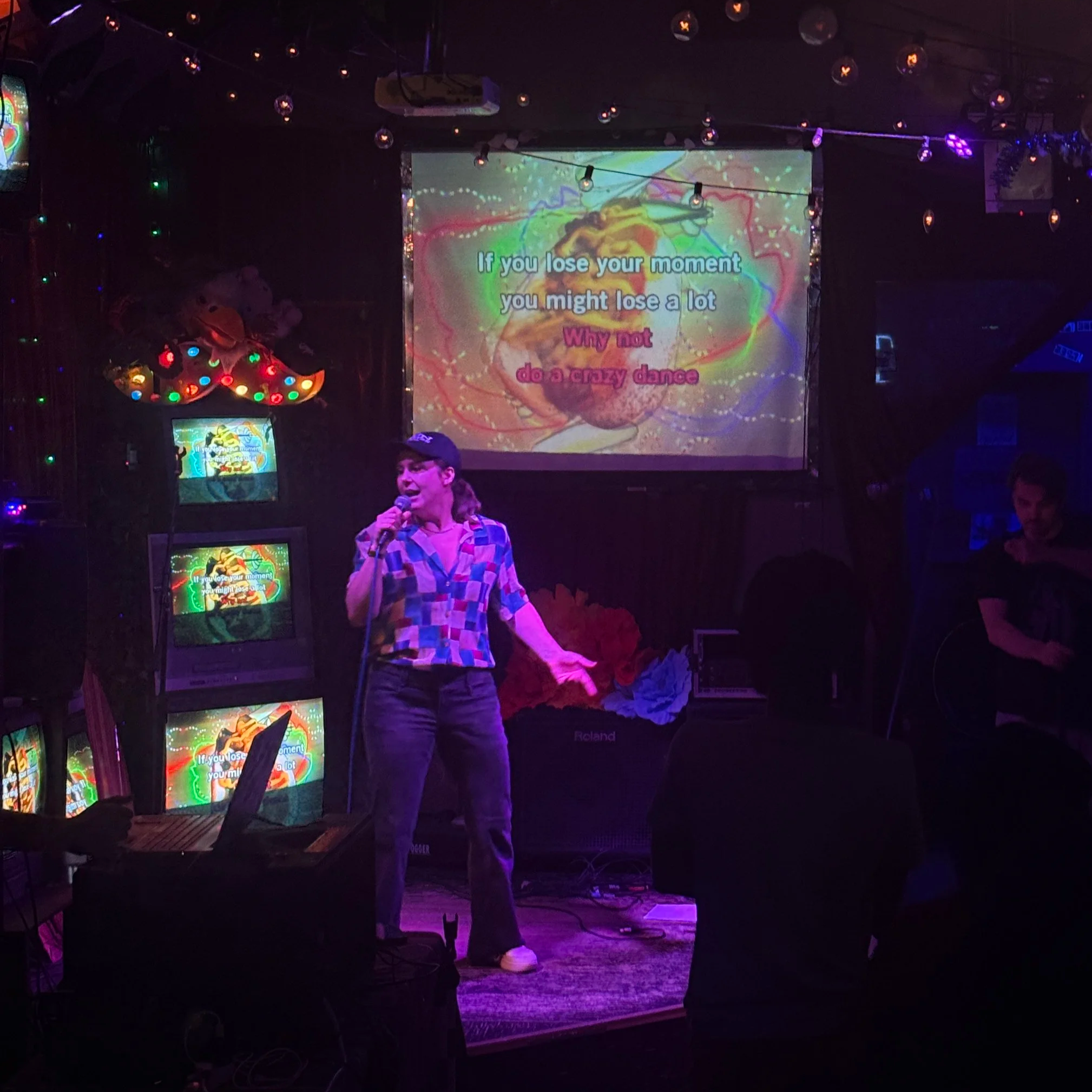 A woman singing on stage with a microphone in a karaoke bar, with a colorful projection on the screen behind her displaying lyrics and art, surrounded by small screens showing the same lyrics.