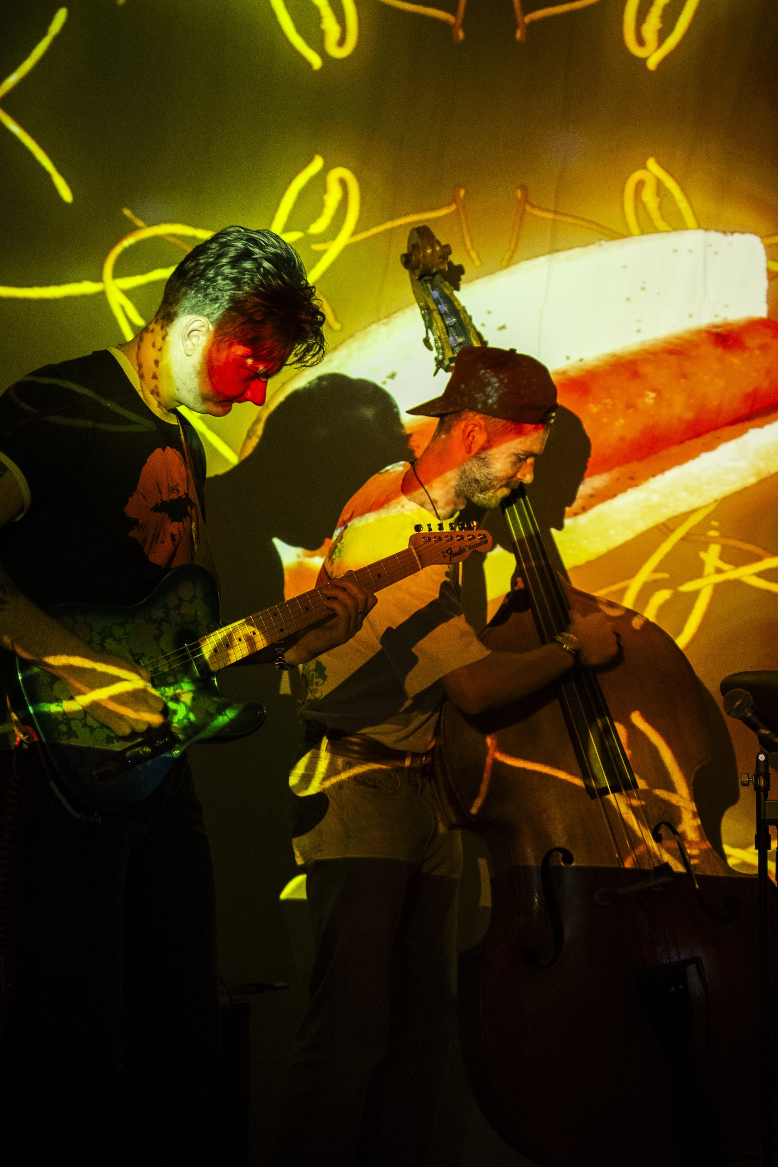 Two musicians playing guitars and a double bass during a live performance, with colorful, abstract visuals projected in the background.