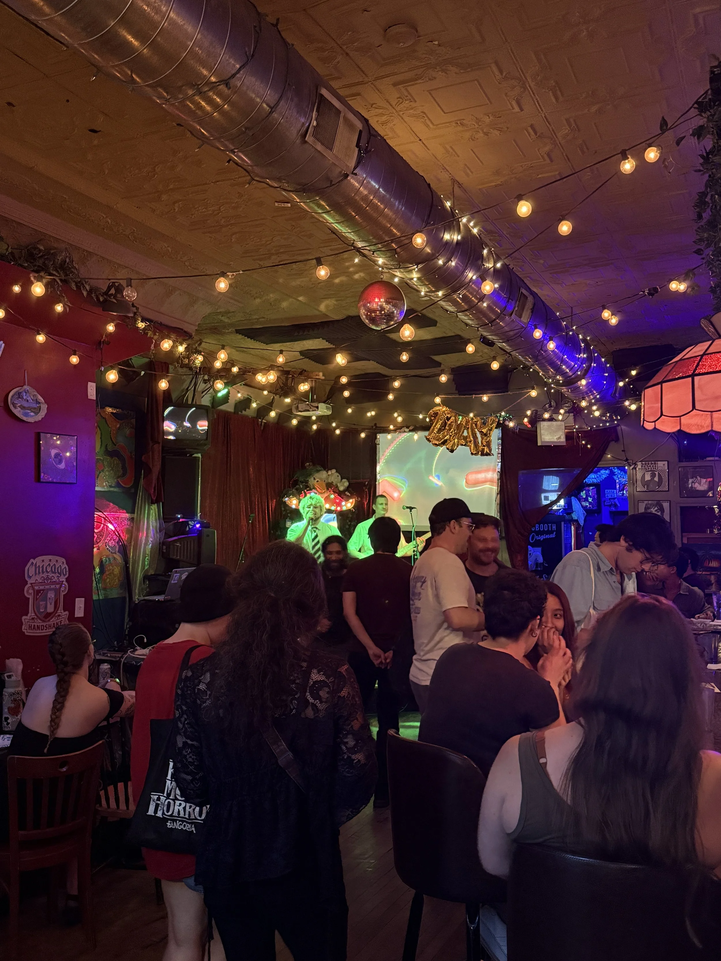 People gathered in a dimly lit bar or music venue with string lights hanging from the ceiling, a disco ball, and a live band performing on stage. The atmosphere is lively and social.