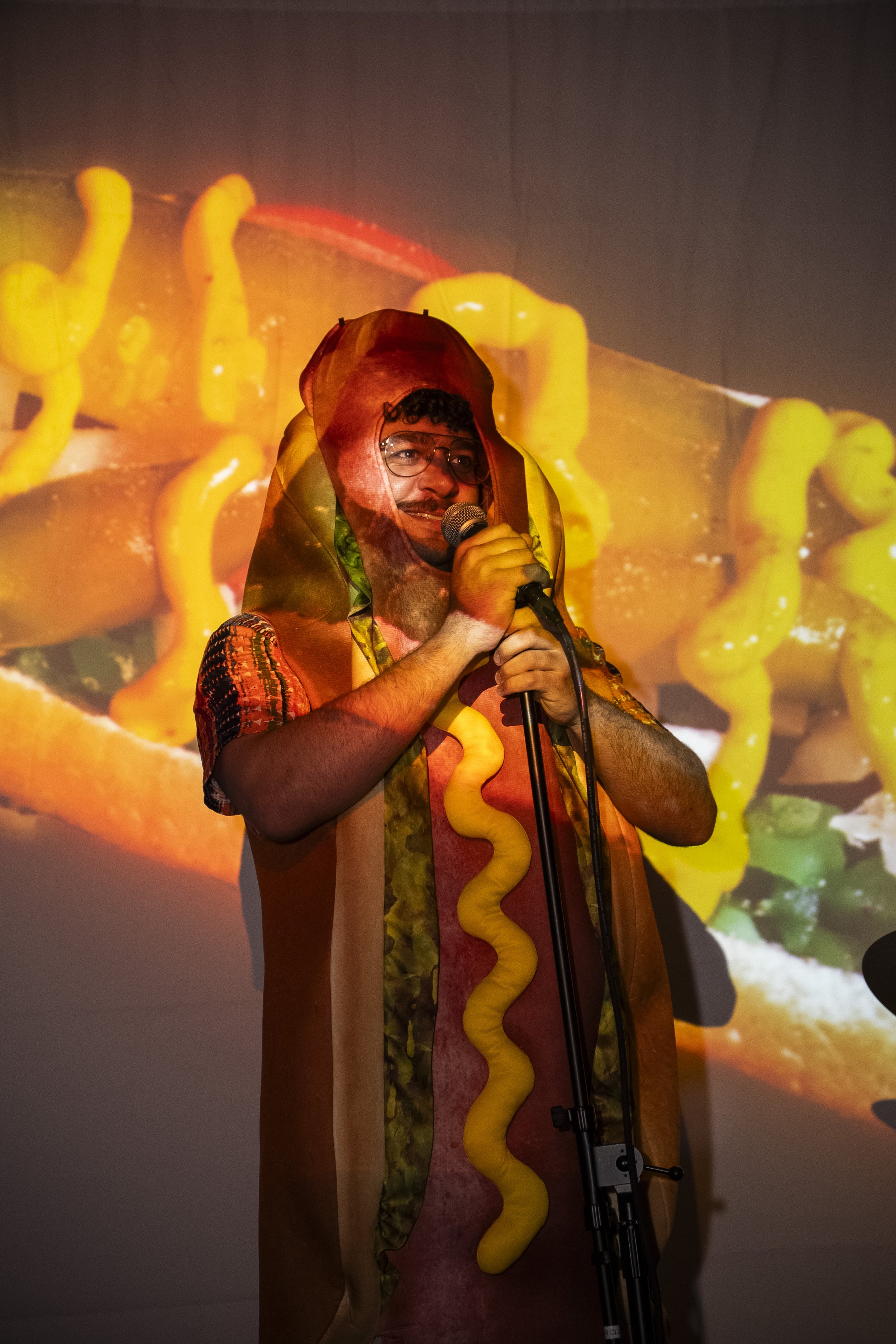 Person wearing hot dog costume with mustard and relish, speaking into a microphone on stage, with a colorful food-themed background projection.