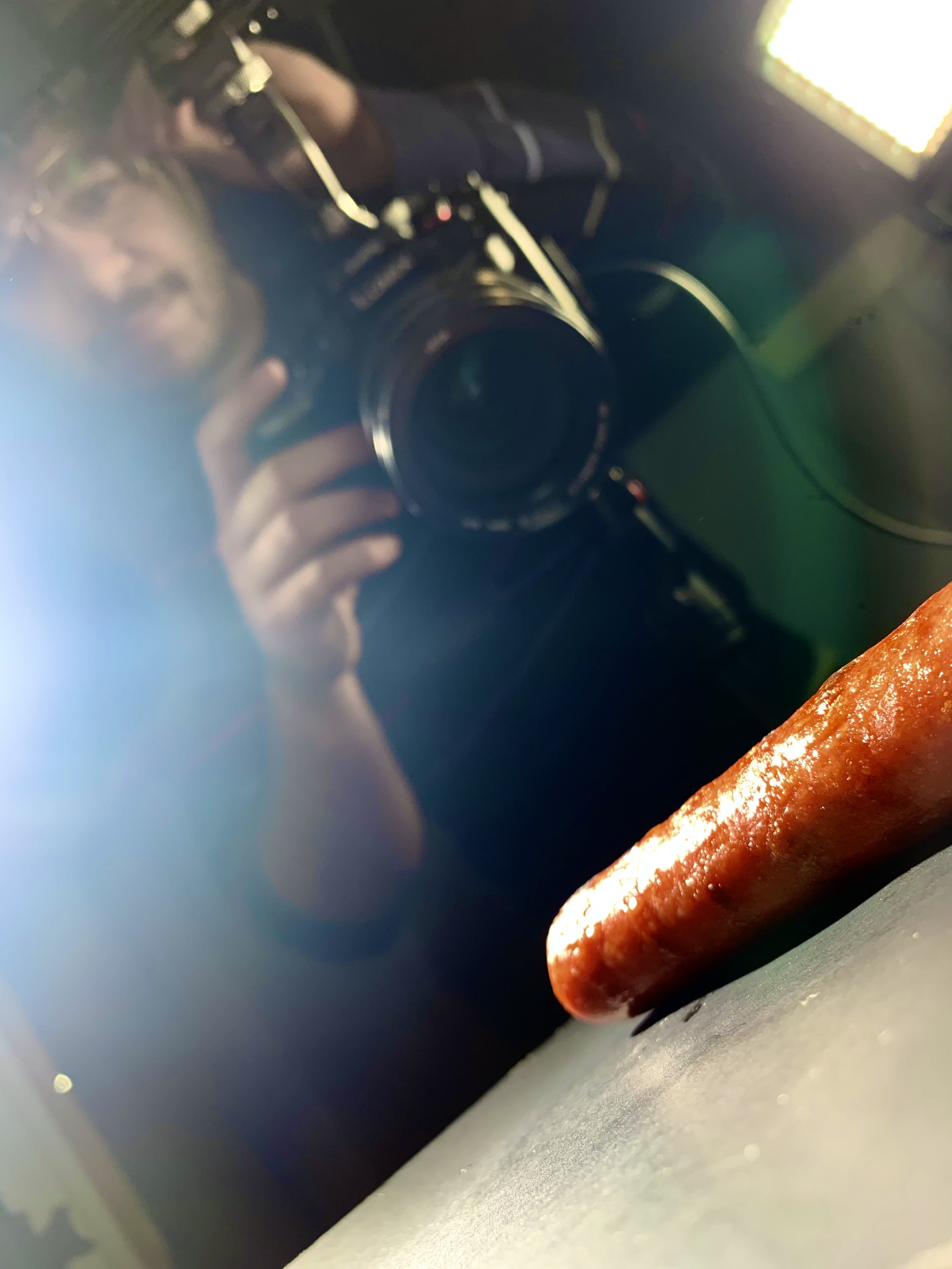 Close-up of a cooked hot dog on a gray surface with a person taking a photo in the background reflected in a mirror.