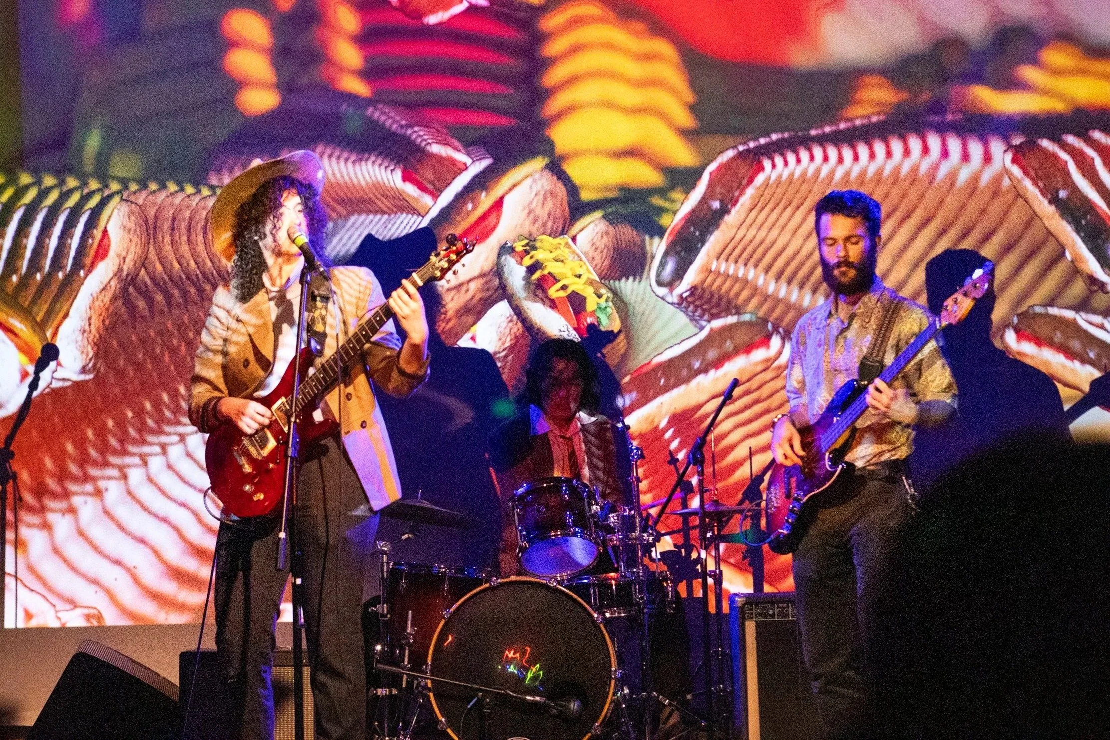 A band of three musicians performing on stage with a colorful background of hot dog ingredients.
