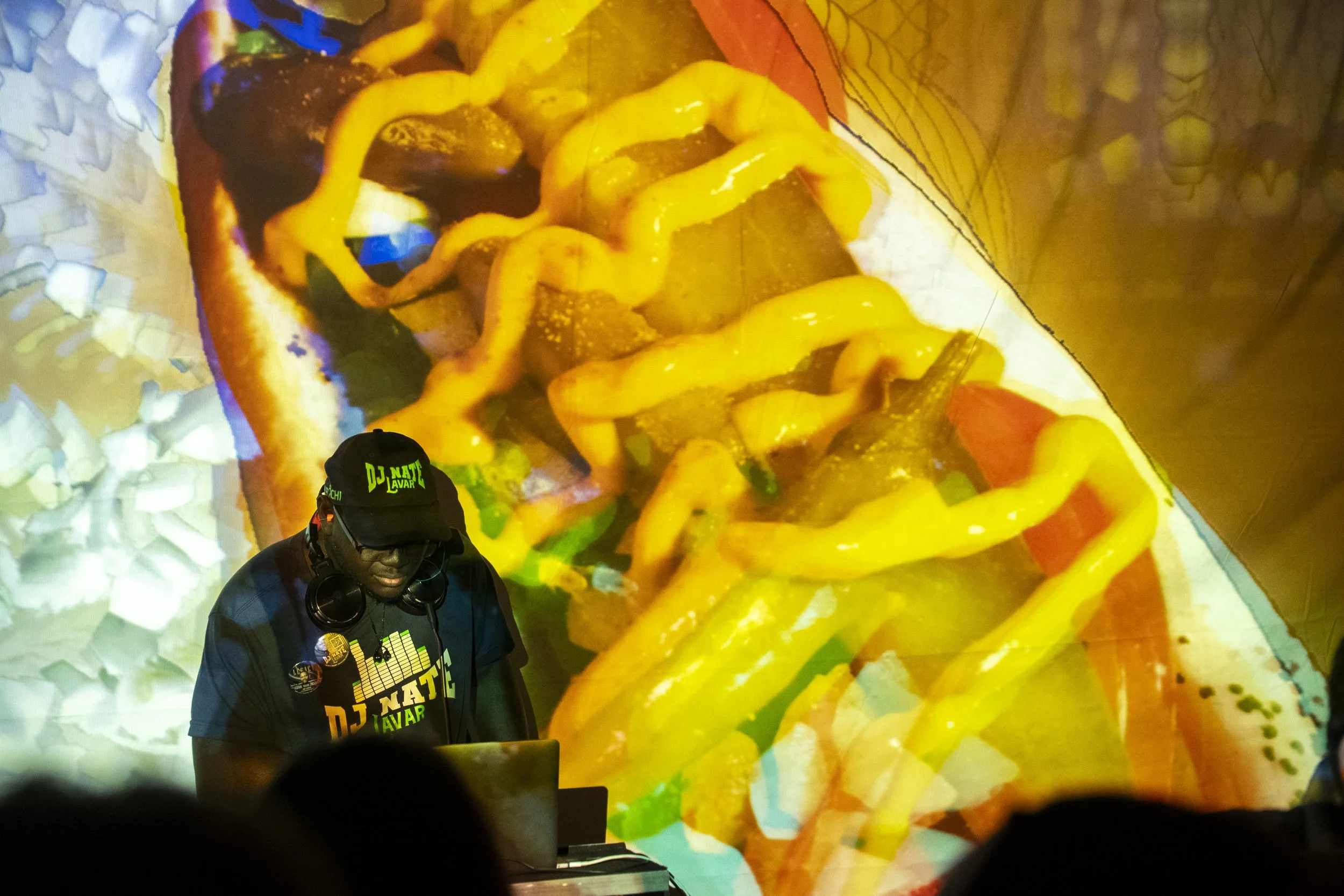 Person DJing with headphones and DJ equipment, wearing a black cap and t-shirt, with large colorful food projection on background.