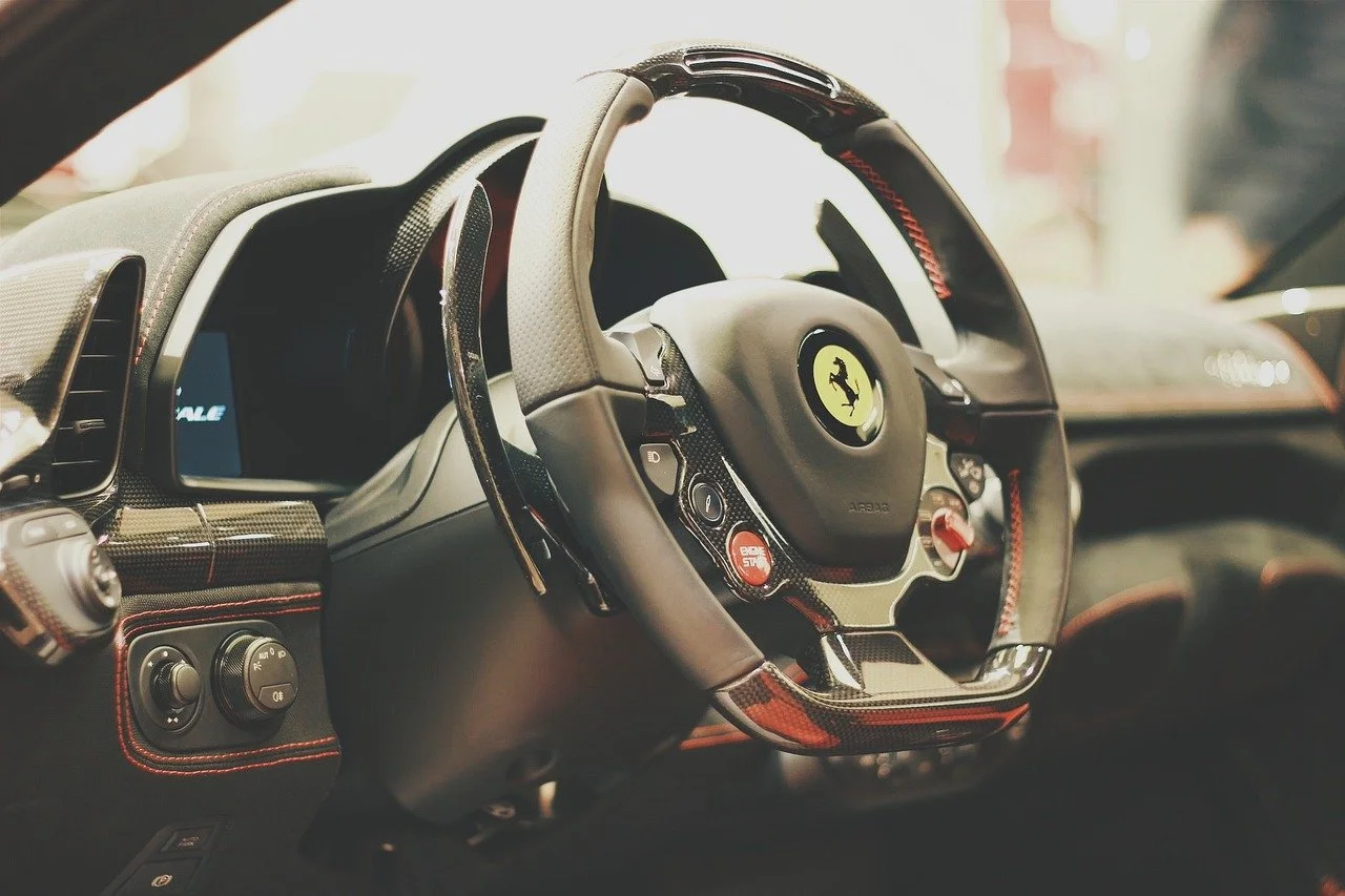 Close-up of a Ferrari steering wheel with black leather and red stitching, featuring the iconic Ferrari logo.