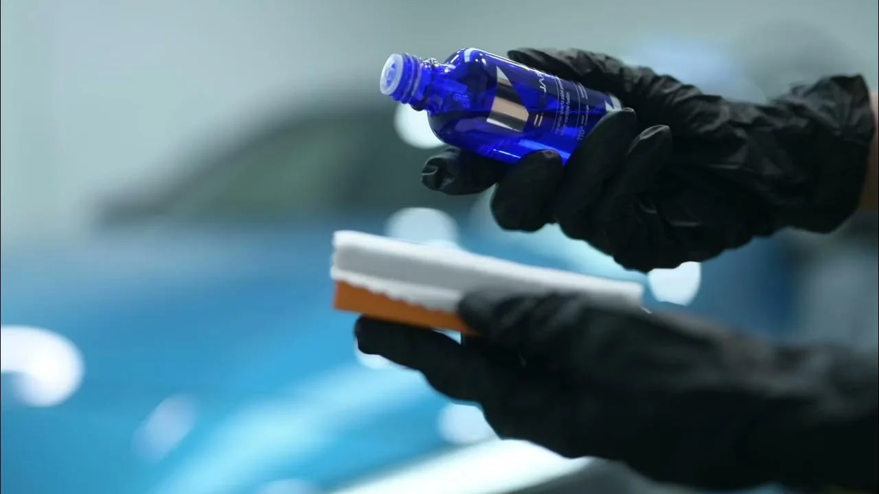 Person wearing black gloves holding a blue bottle and an applicator pad.