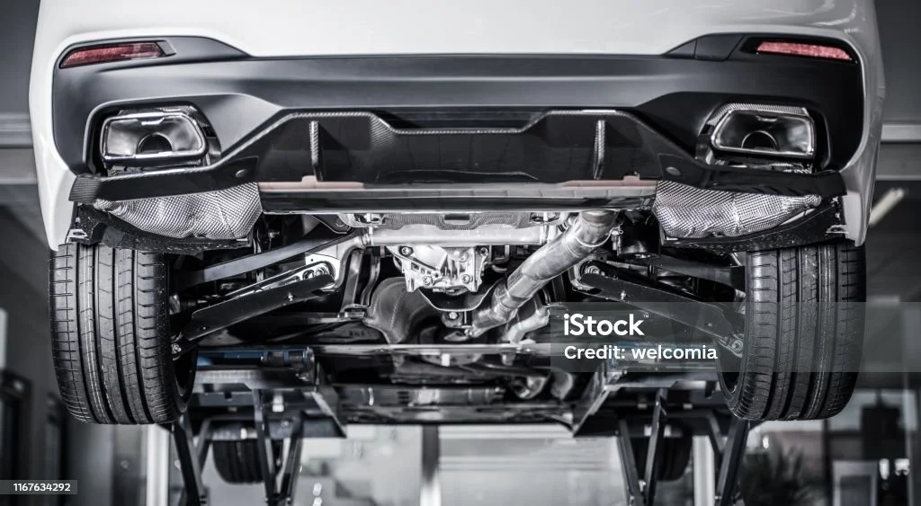 Underside of a car showing suspension, exhaust system, and rear wheels, elevated on a lift in an auto repair shop.