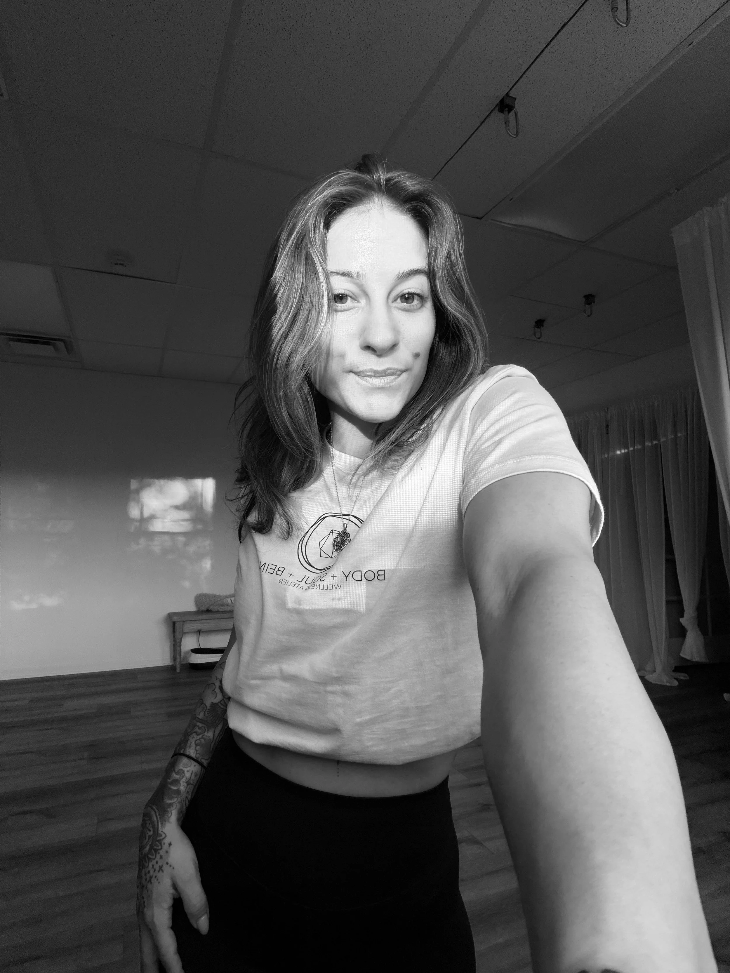 A smiling woman with tattoos in a black and white photo with black pants practicing yoga in a cross-legged seated meditation pose on a mat indoors.