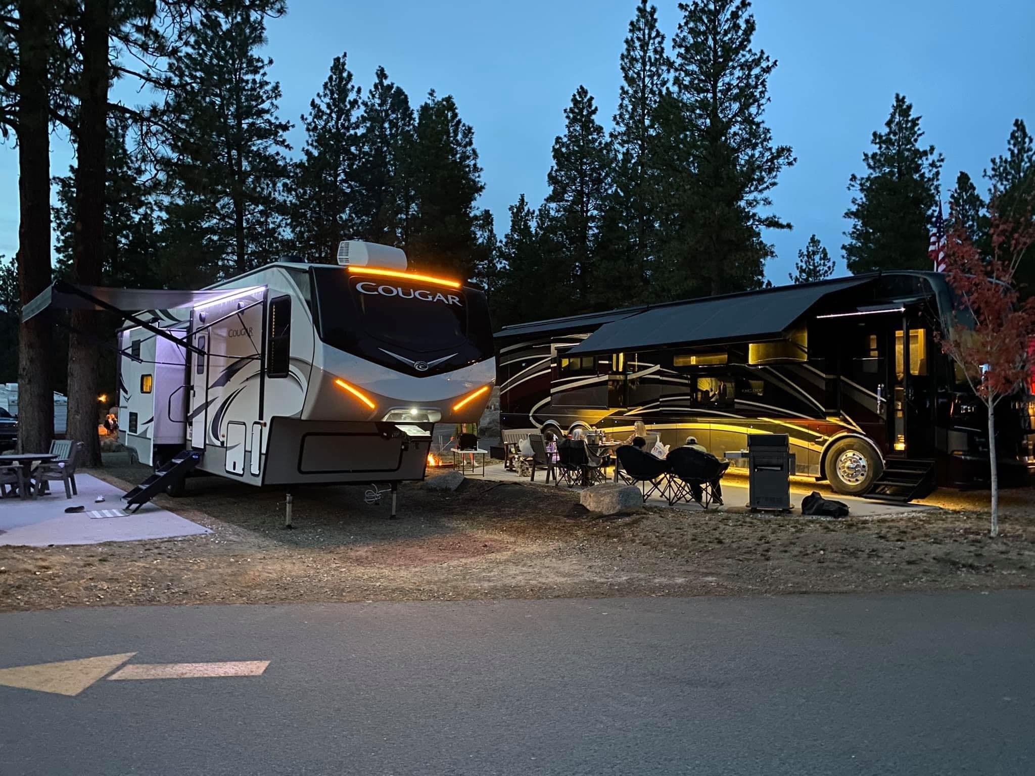 Evening campsite with a Cougar fifth-wheel RV and a luxury motorhome, both illuminated. A group relaxes by a fire pit under tall pine trees.