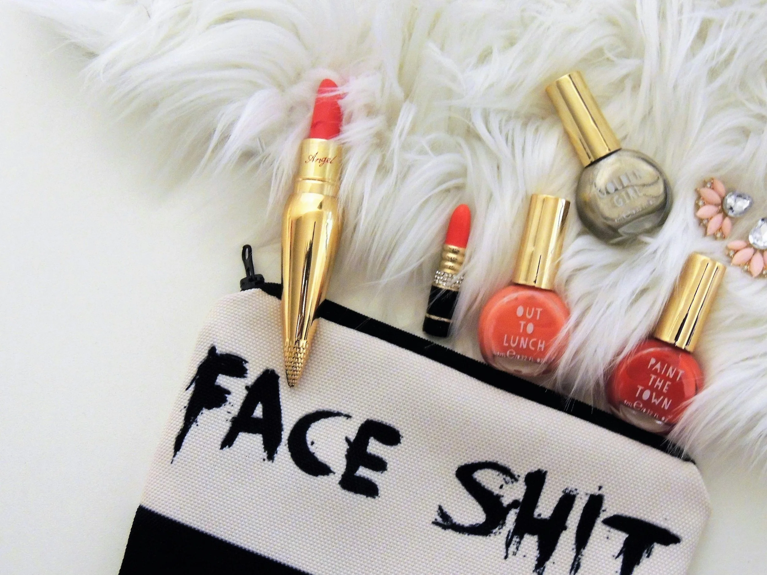 A flat lay of cosmetics and accessories on a white faux fur surface, including a gold-tubed red lipstick, a small black and gold red lipstick, three nail polishes with gold caps, a pair of earrings with pink and clear stones, and a makeup bag with text "FACE SHIT."