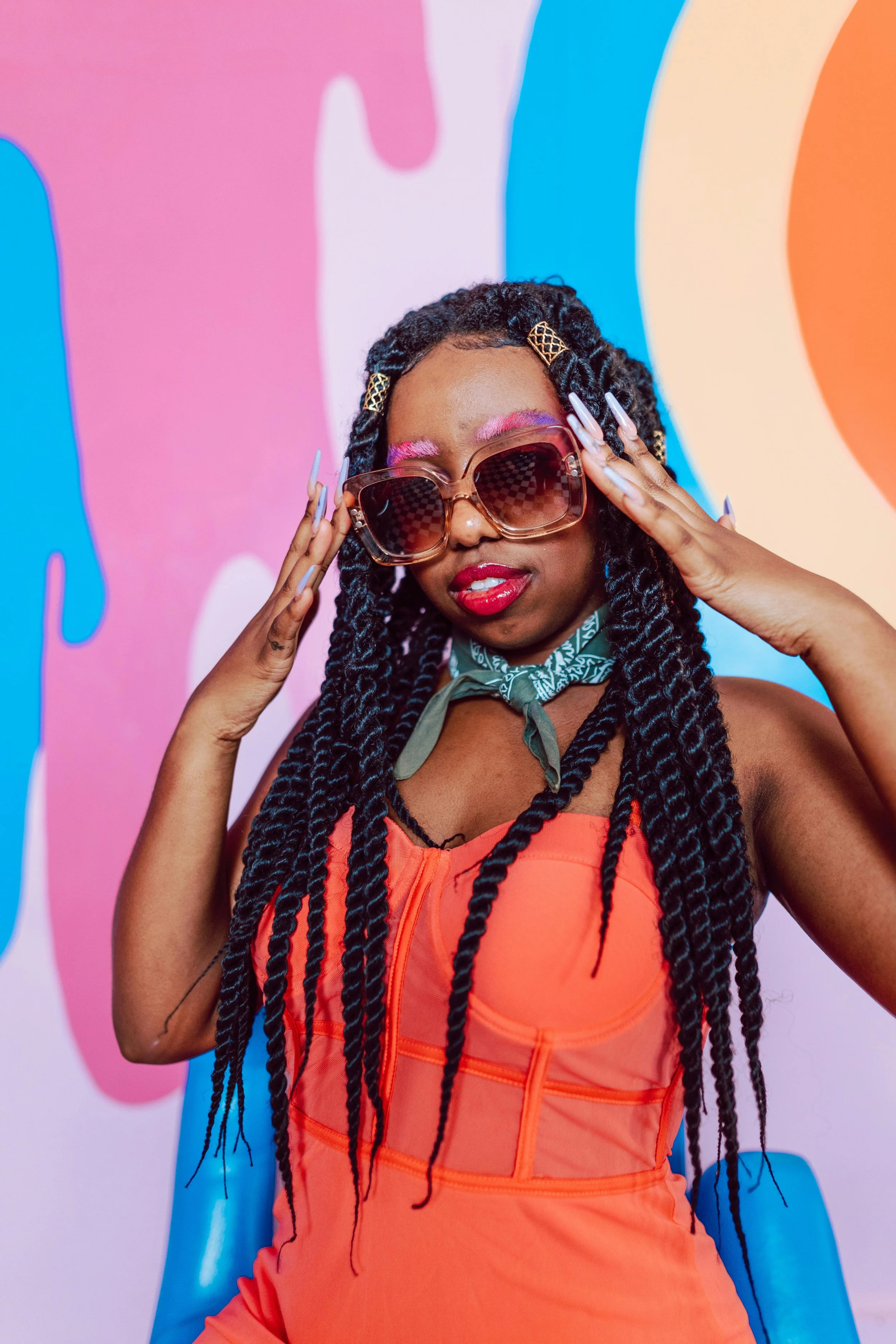 Person with long braids wearing large sunglasses, an orange outfit, and a green scarf, posing in front of a colorful abstract background.