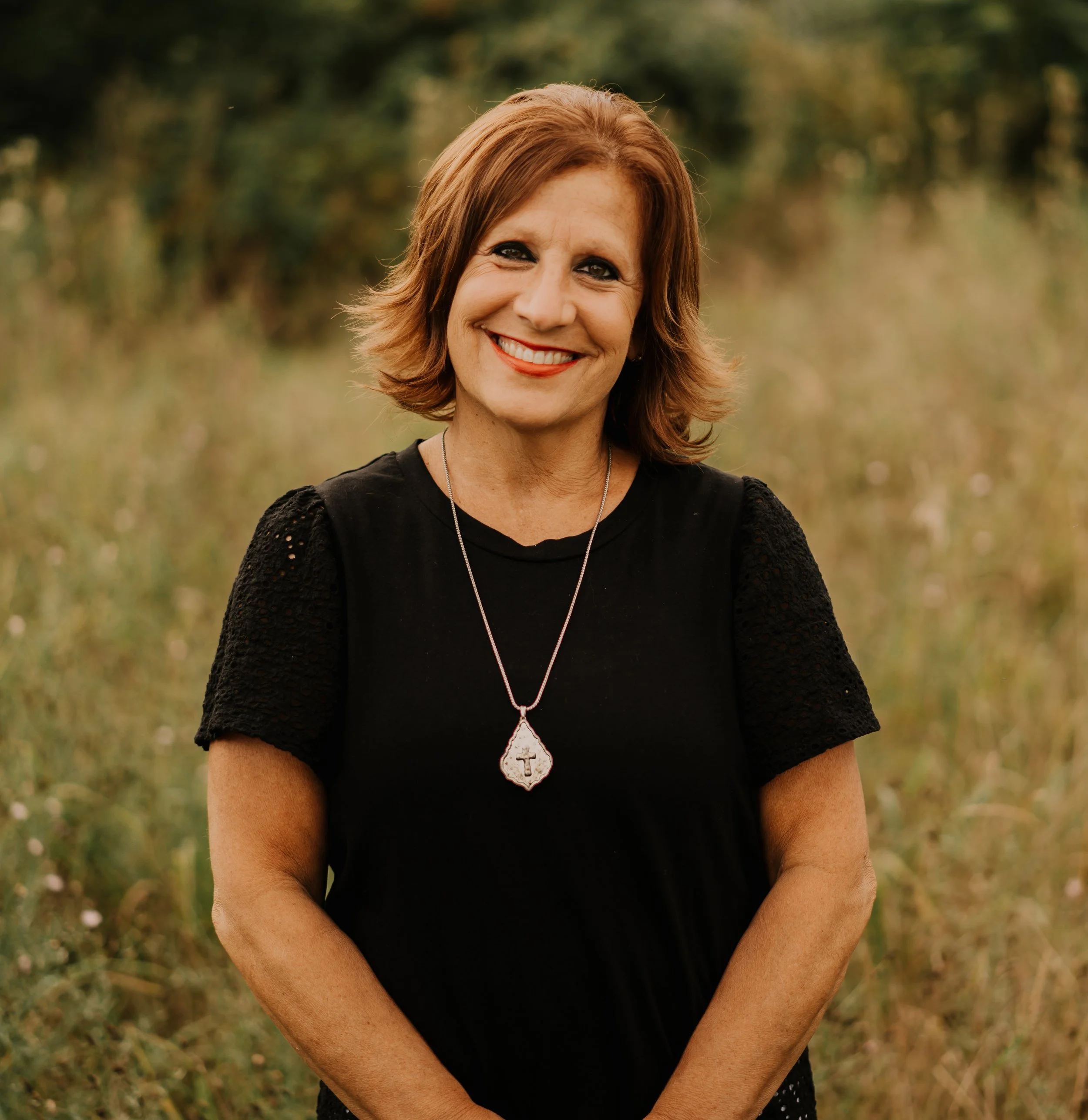 A smiling woman with shoulder-length reddish-brown hair wearing a black top with textured sleeves and a pendant necklace, standing outdoors in a grassy field with trees in the background.