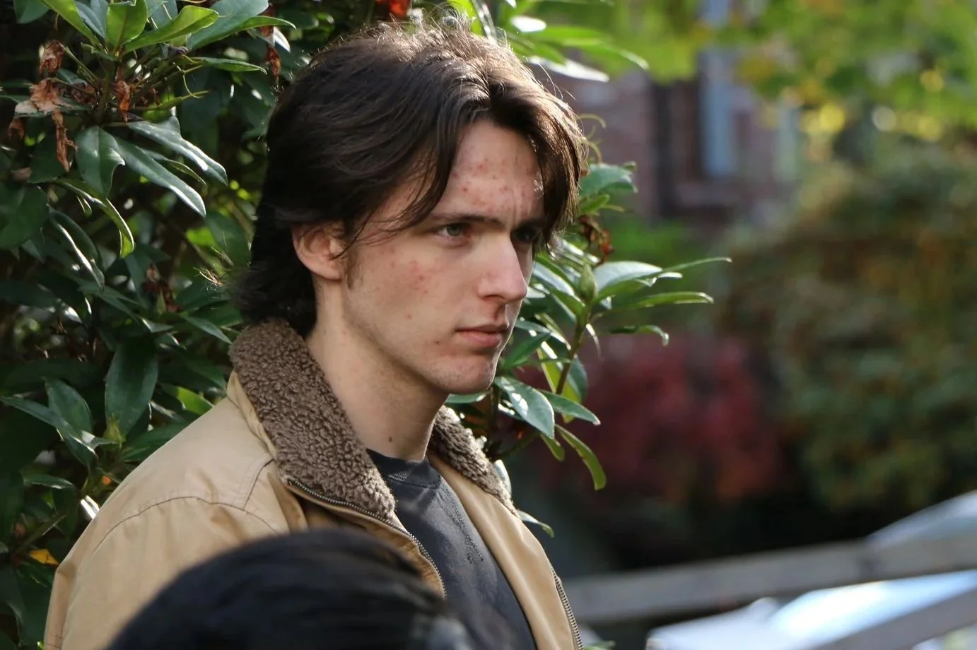 A young man with dark brown hair, light skin, and acne on his face, wearing a tan jacket with a fur collar, standing outdoors near green leafy bushes.