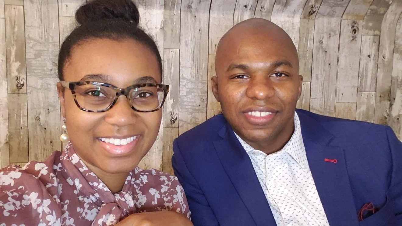 A woman with glasses and a bun hairstyle smiling, and a man in a blue blazer smiling, sitting together in front of a wooden wall.