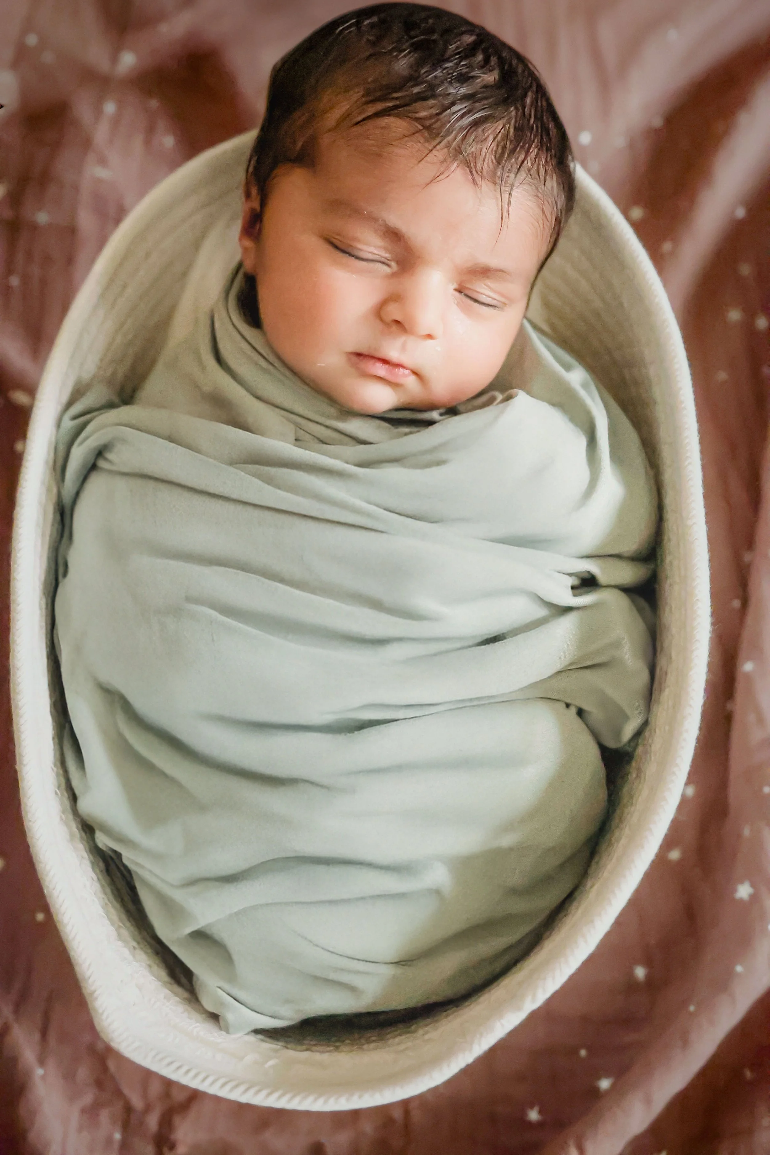 A sleeping baby wrapped in a light green blanket, lying in a woven basket.