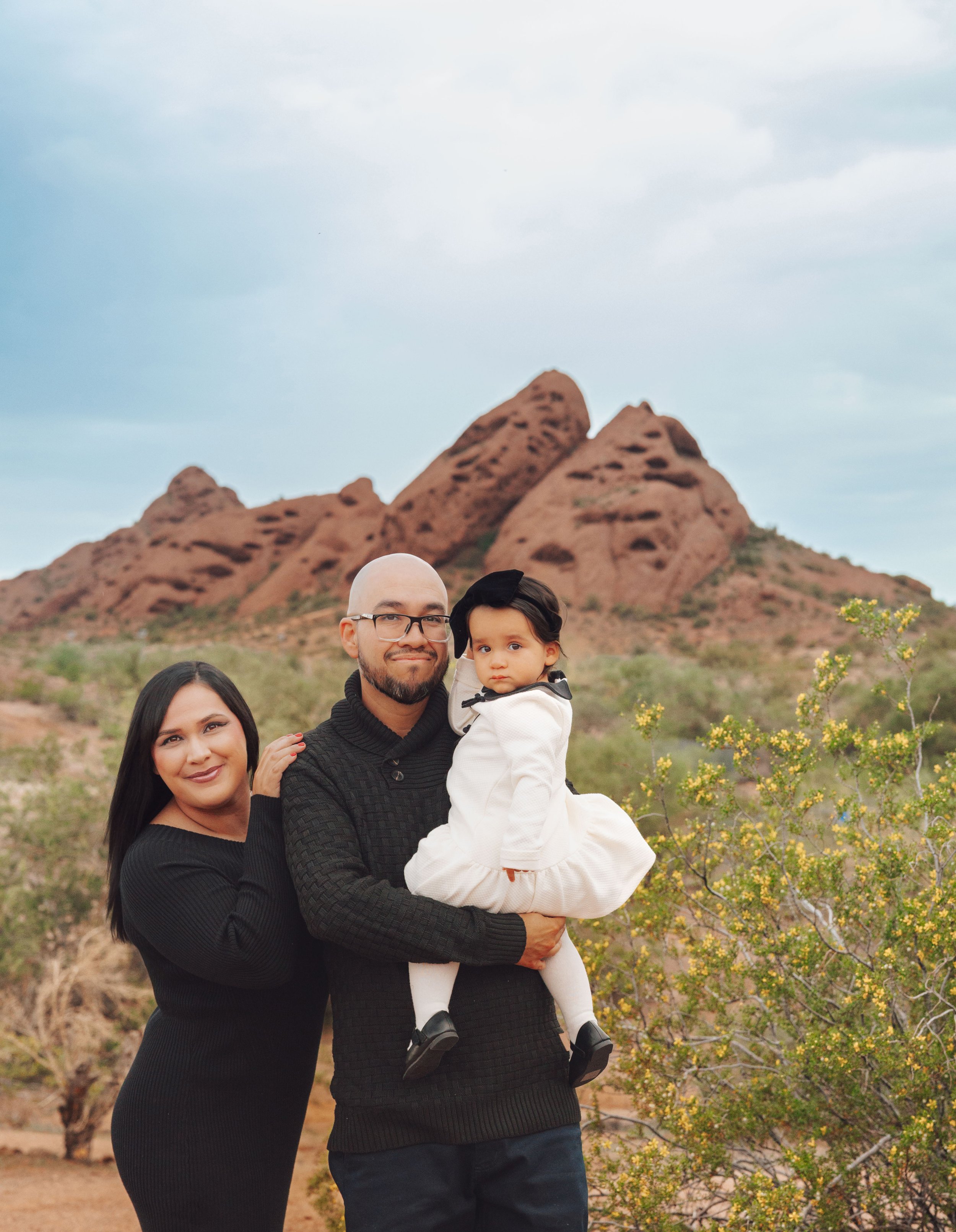 A family of three standing outdoors with red rock formations in the background, shrubs, and yellow flowering plants.