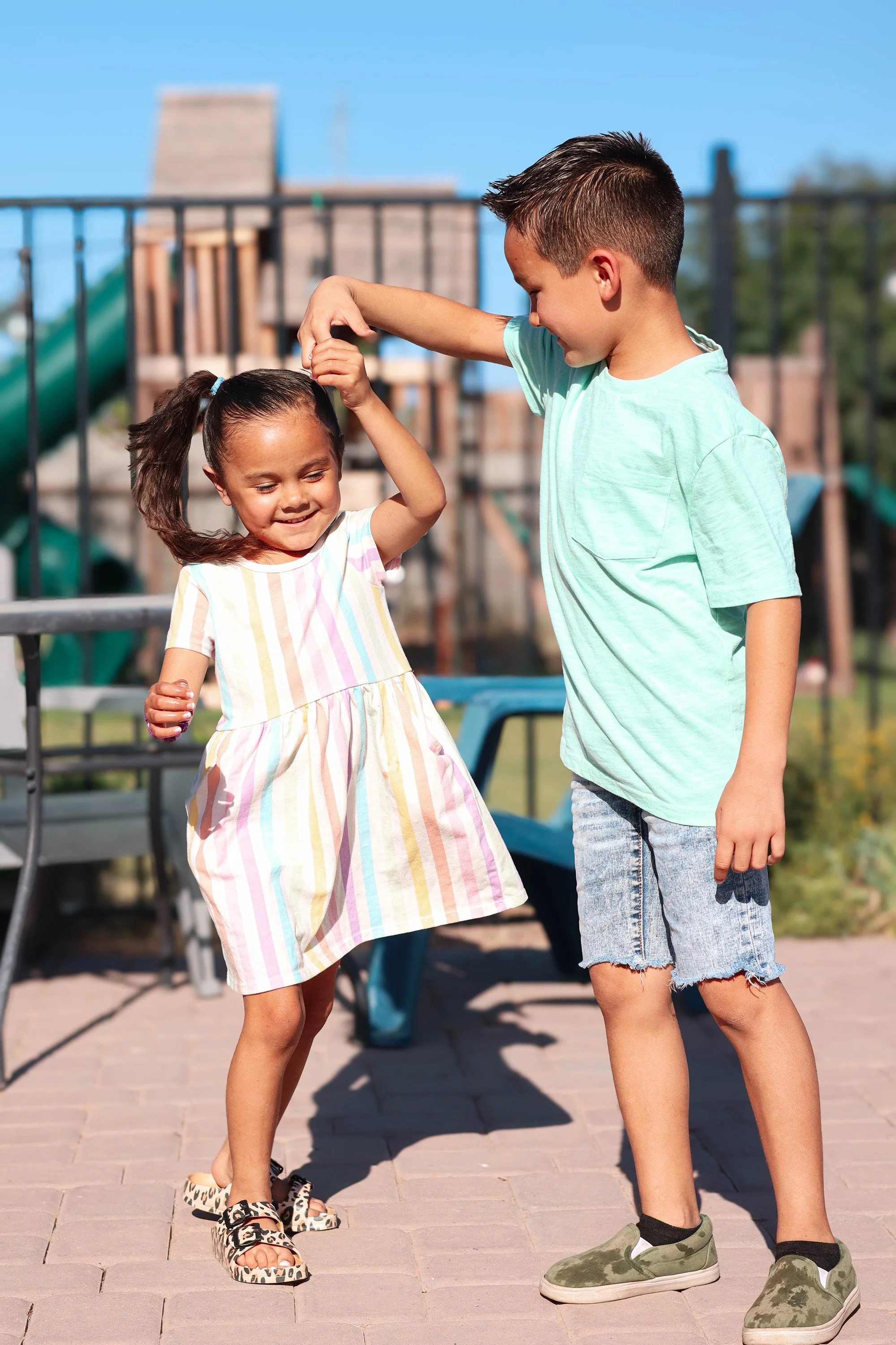 A boy and girl play-fight on a playground on a sunny day, with the boy pretending to pull the girl's hair while both are smiling.