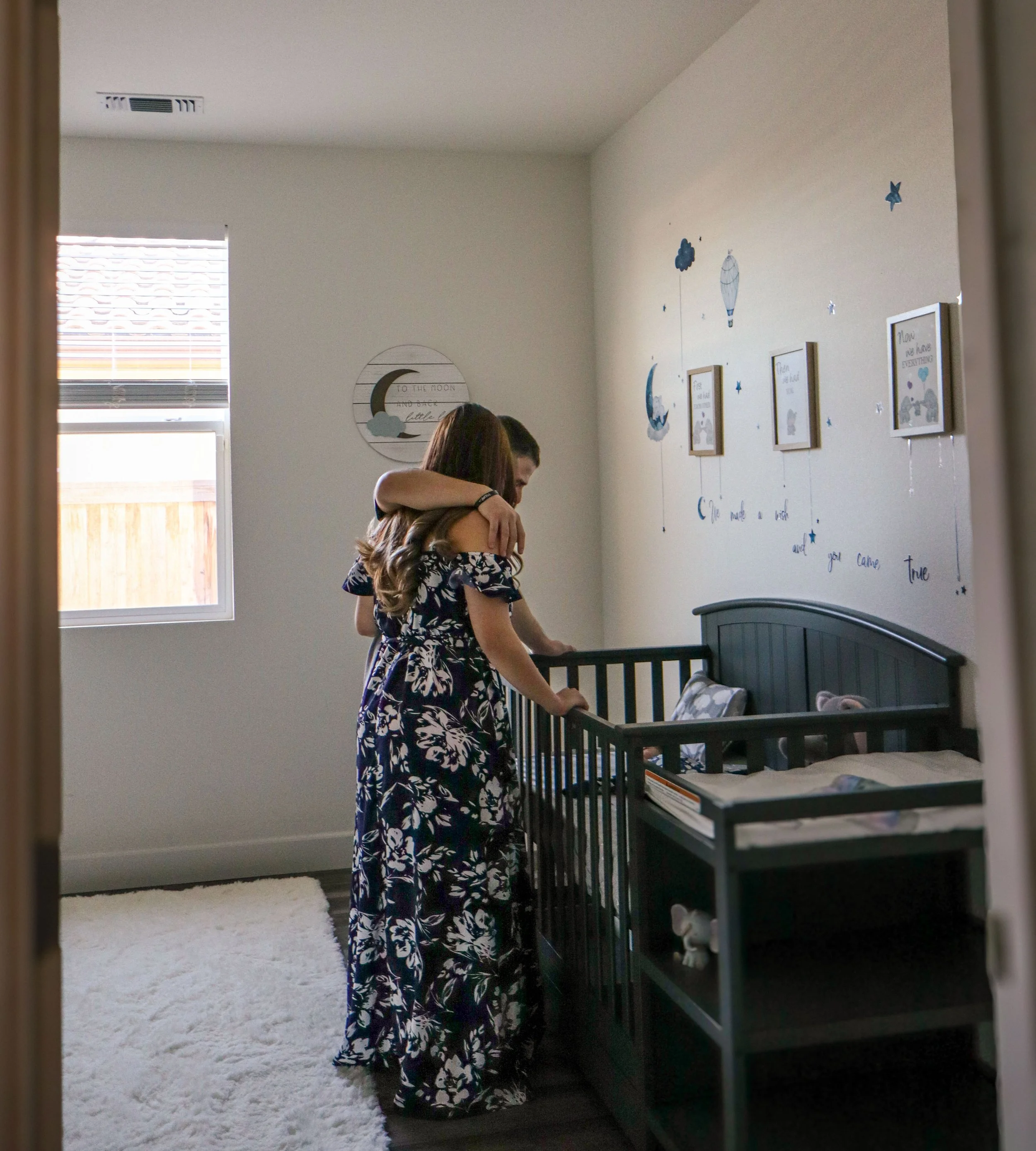 A woman and a young girl hugging in a nursery with a crib, wall decorations, and a window.