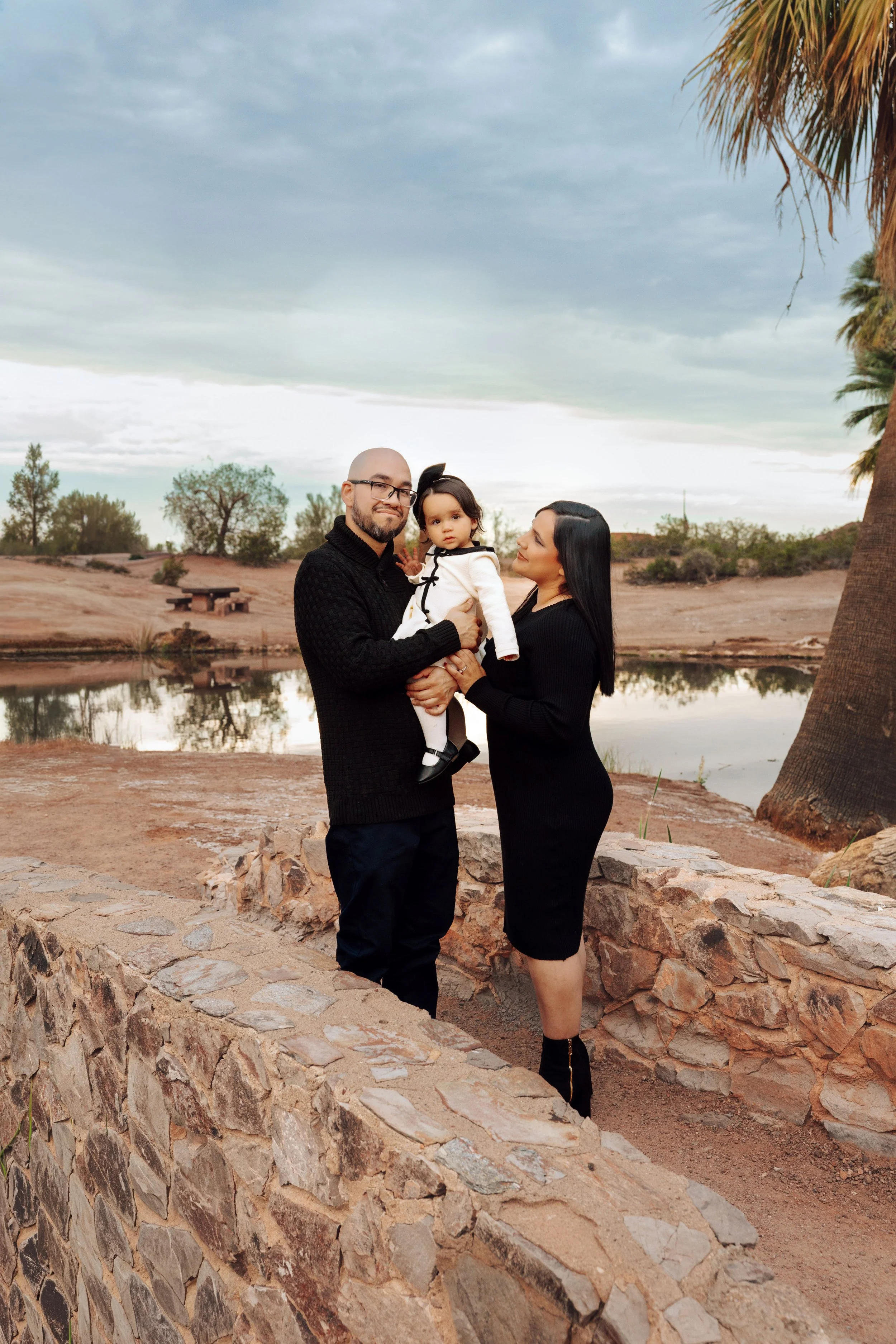 A family of three standing outdoors near a small lake, with a man holding a young girl and a woman standing next to them, all dressed in black and white, with trees and cloudy sky in the background.