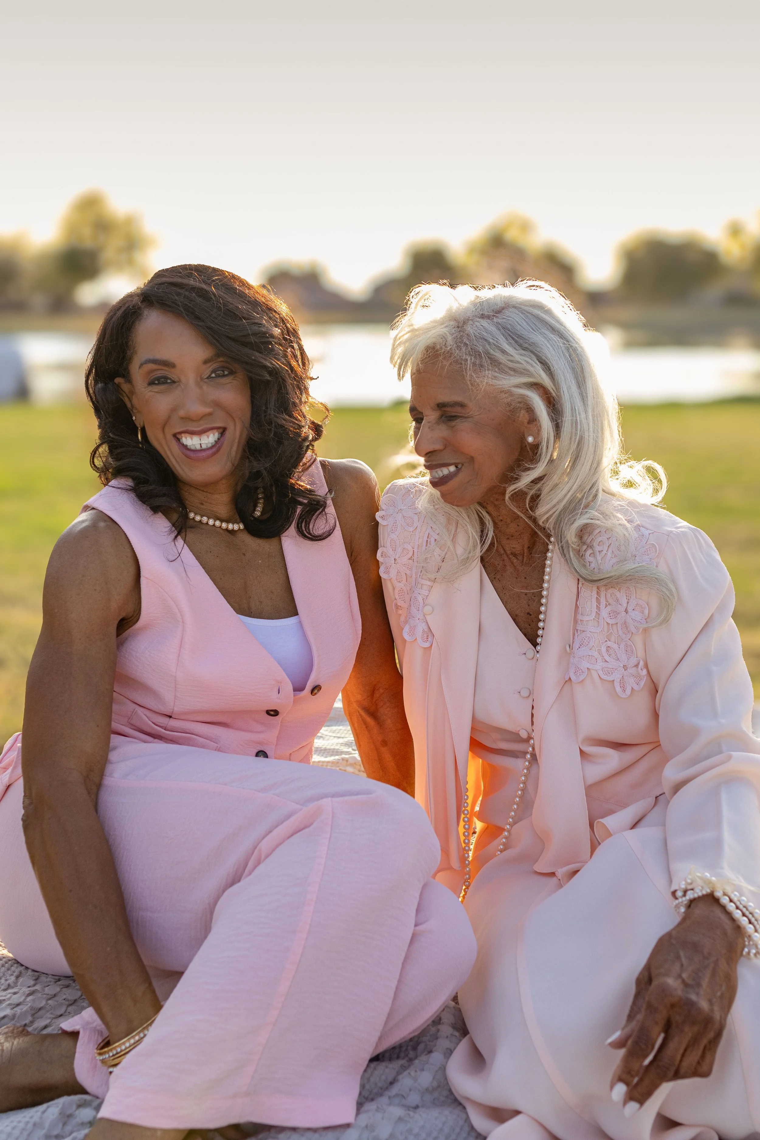 Two women sitting outdoors on a grassy area near a body of water, enjoying each other's company at sunset. They are smiling and dressed in light pink and cream clothing, wearing pearl jewelry.