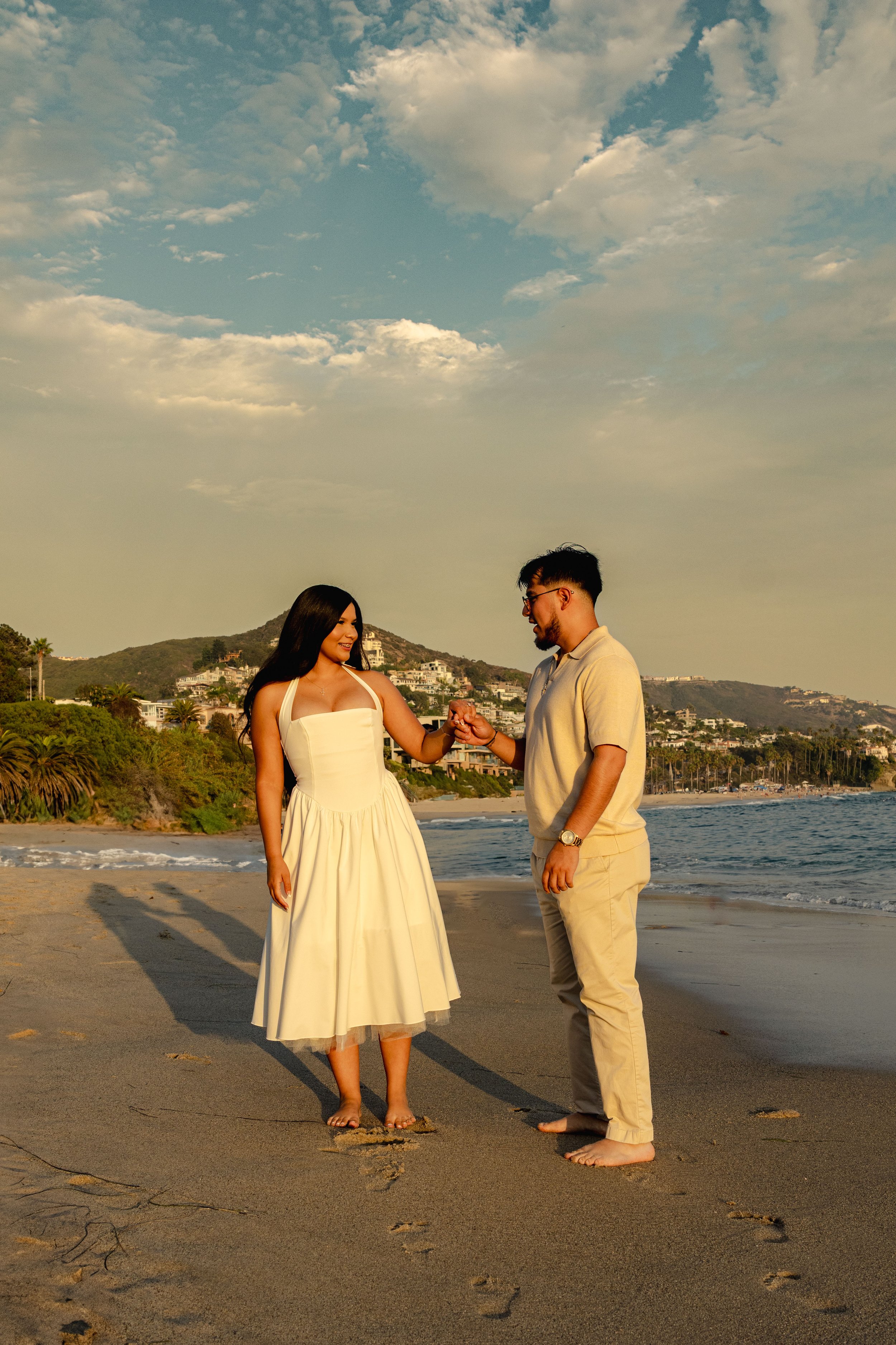 A man and woman holding hands on a beach during sunset, with hills and houses in the background.
