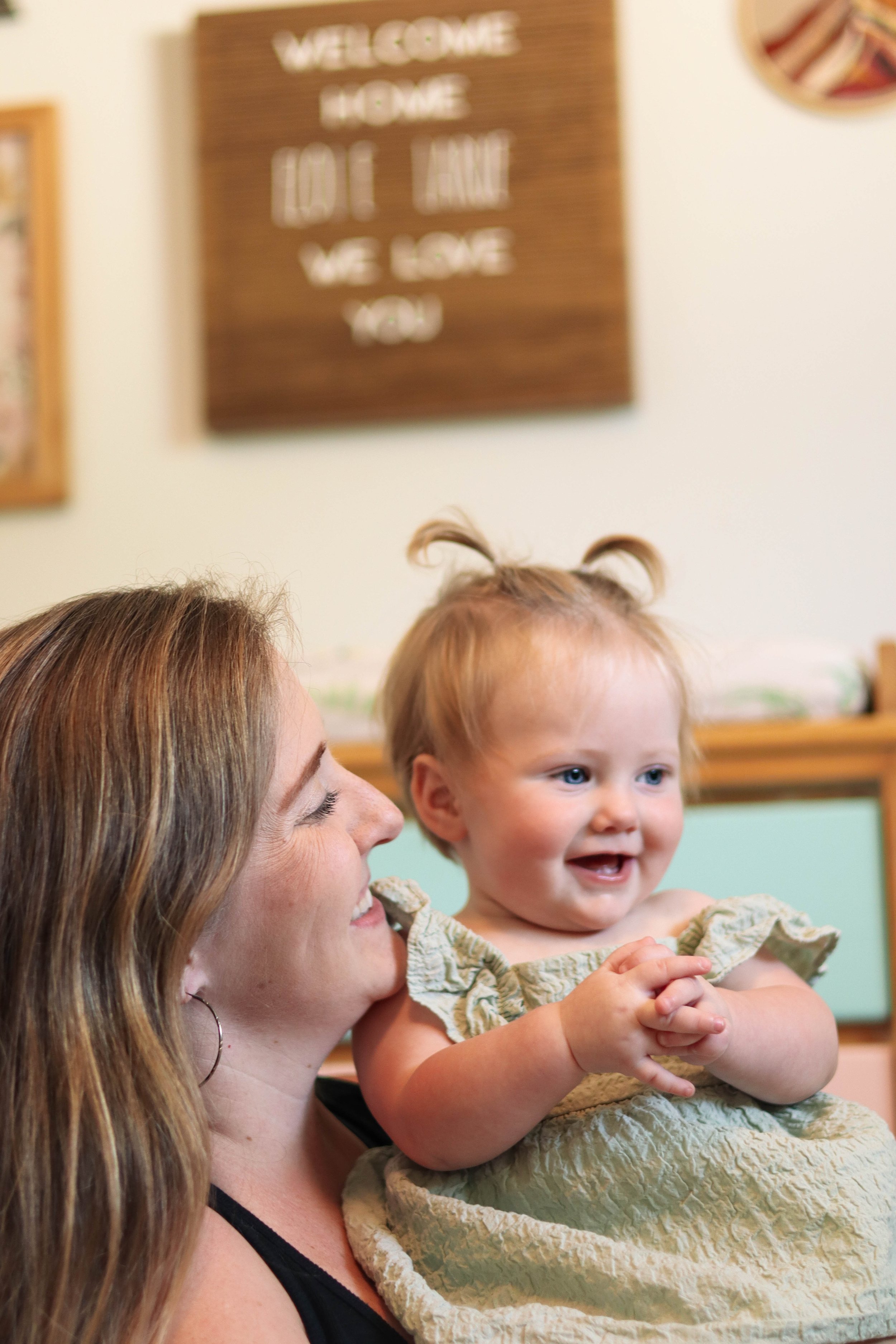 A woman holding a smiling baby girl with curly hair in a yellow dress, living room background