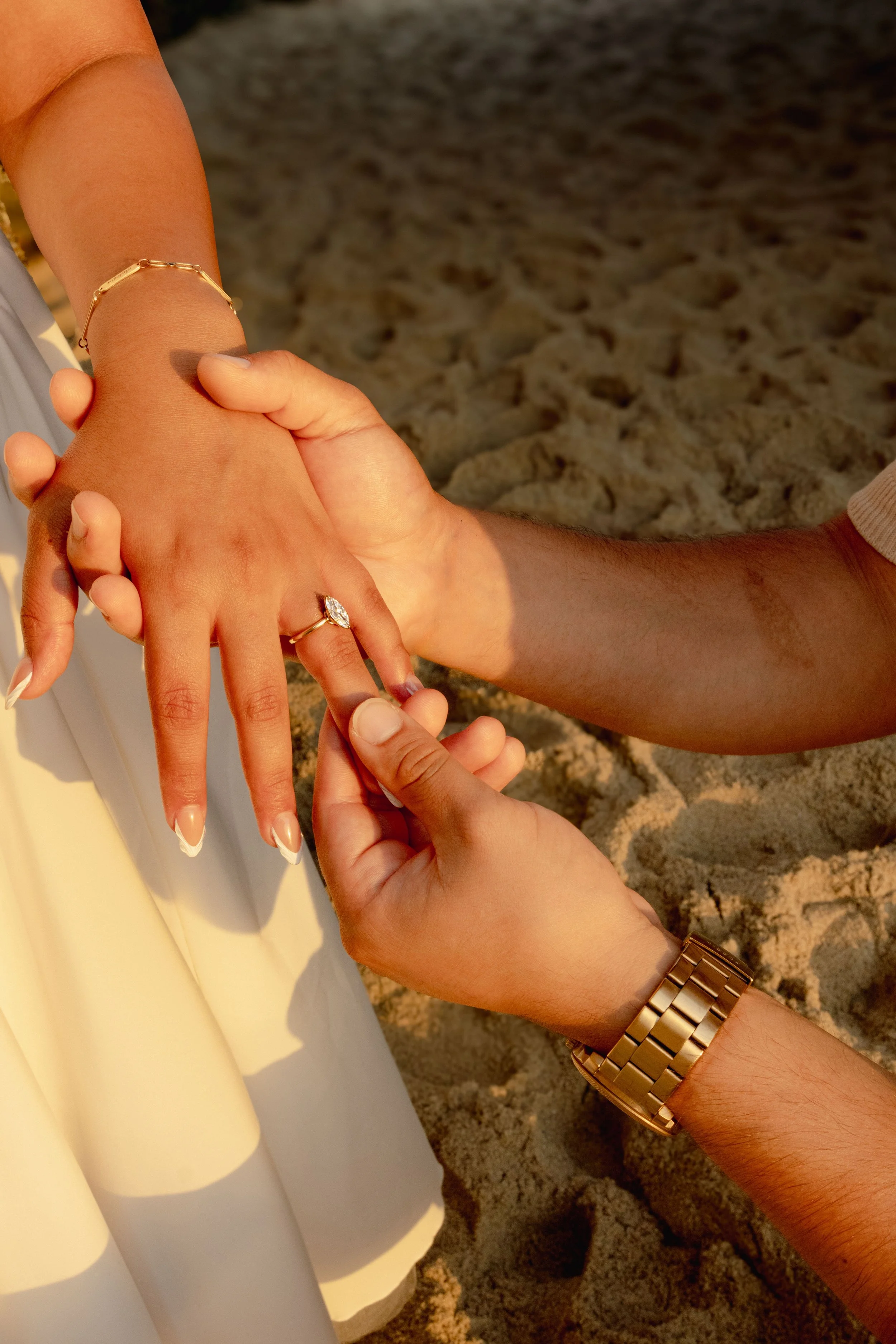 A person is having their ring finger with an engagement ring held by another person, on a sandy beach under warm sunlight.