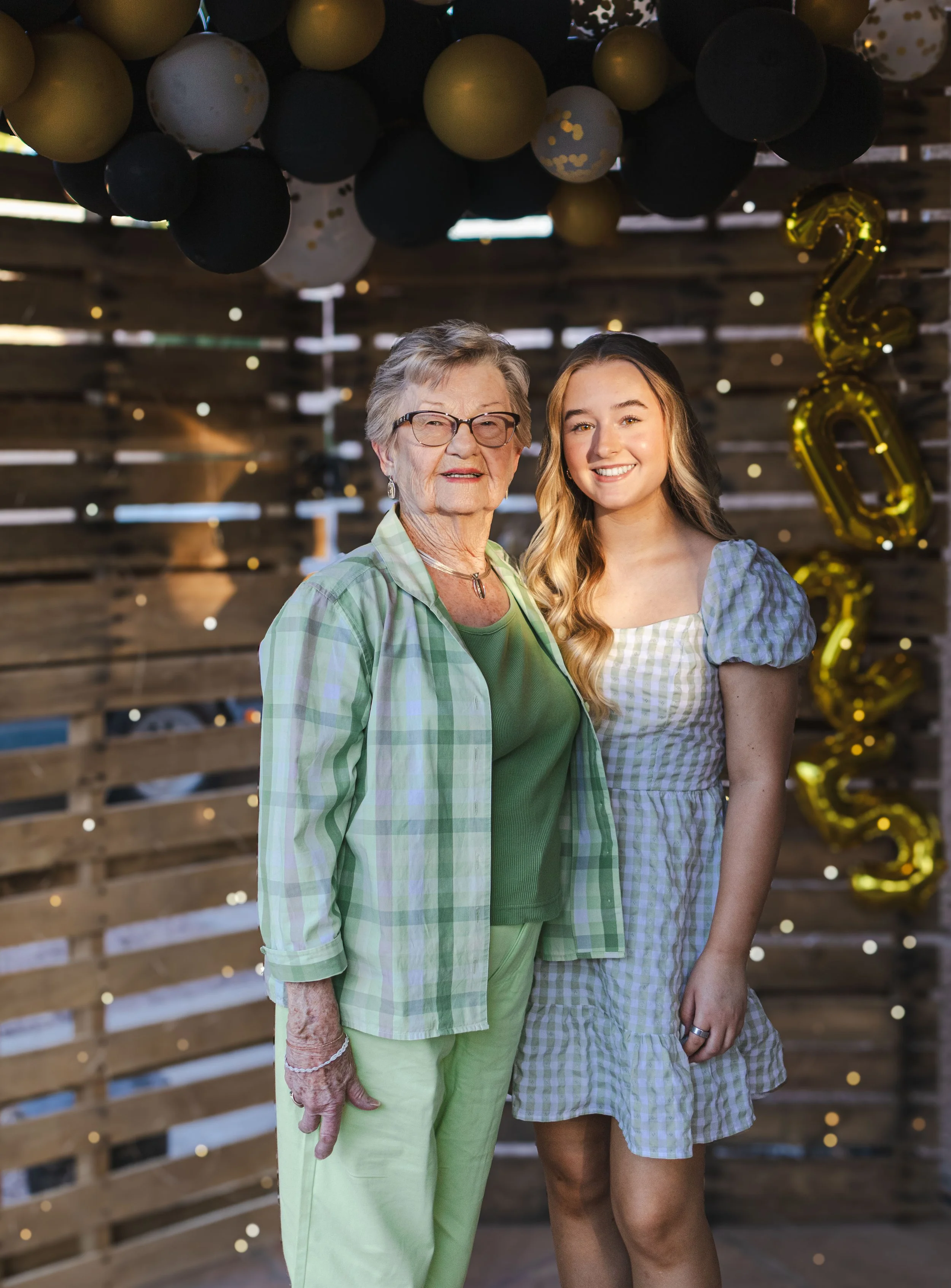 Two women smiling at a celebration with black, gold, and white balloons and gold '2023' balloons in the background.