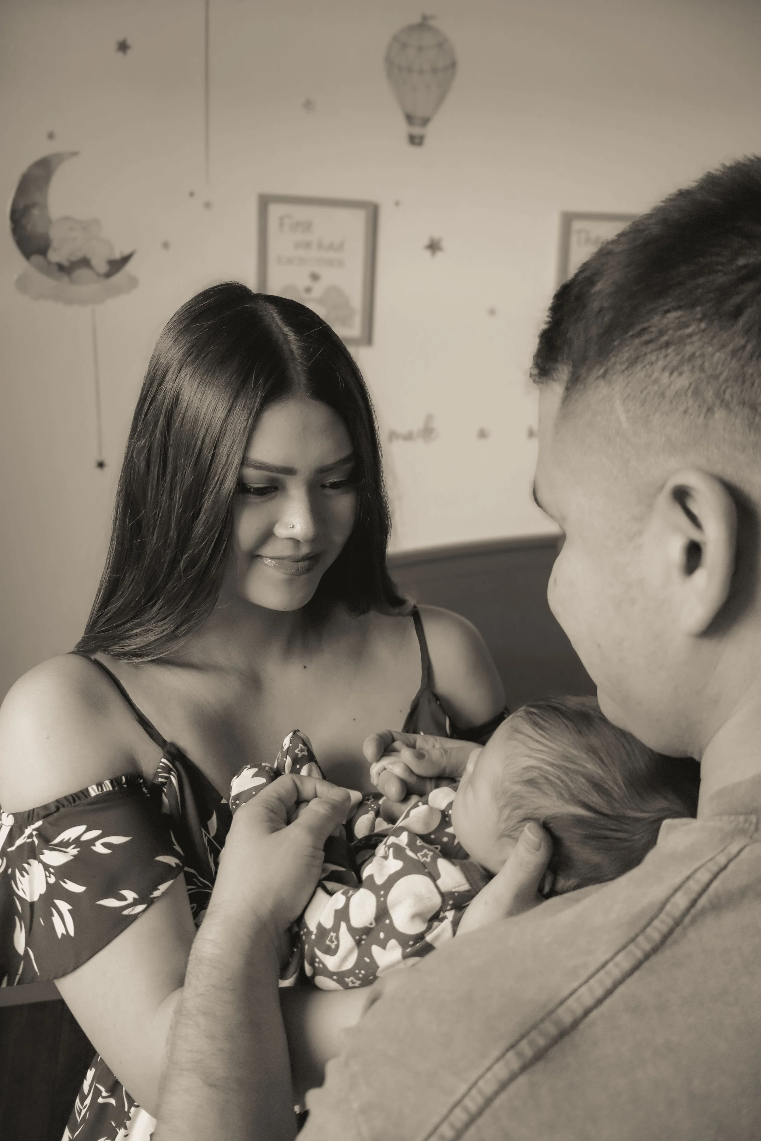A woman with long dark hair and a nose piercing looks at a newborn baby who is being held by a man. The baby is wrapped in a blanket with a pattern of hearts and stars. The background has pastel-colored decorations and framed pictures on the wall.