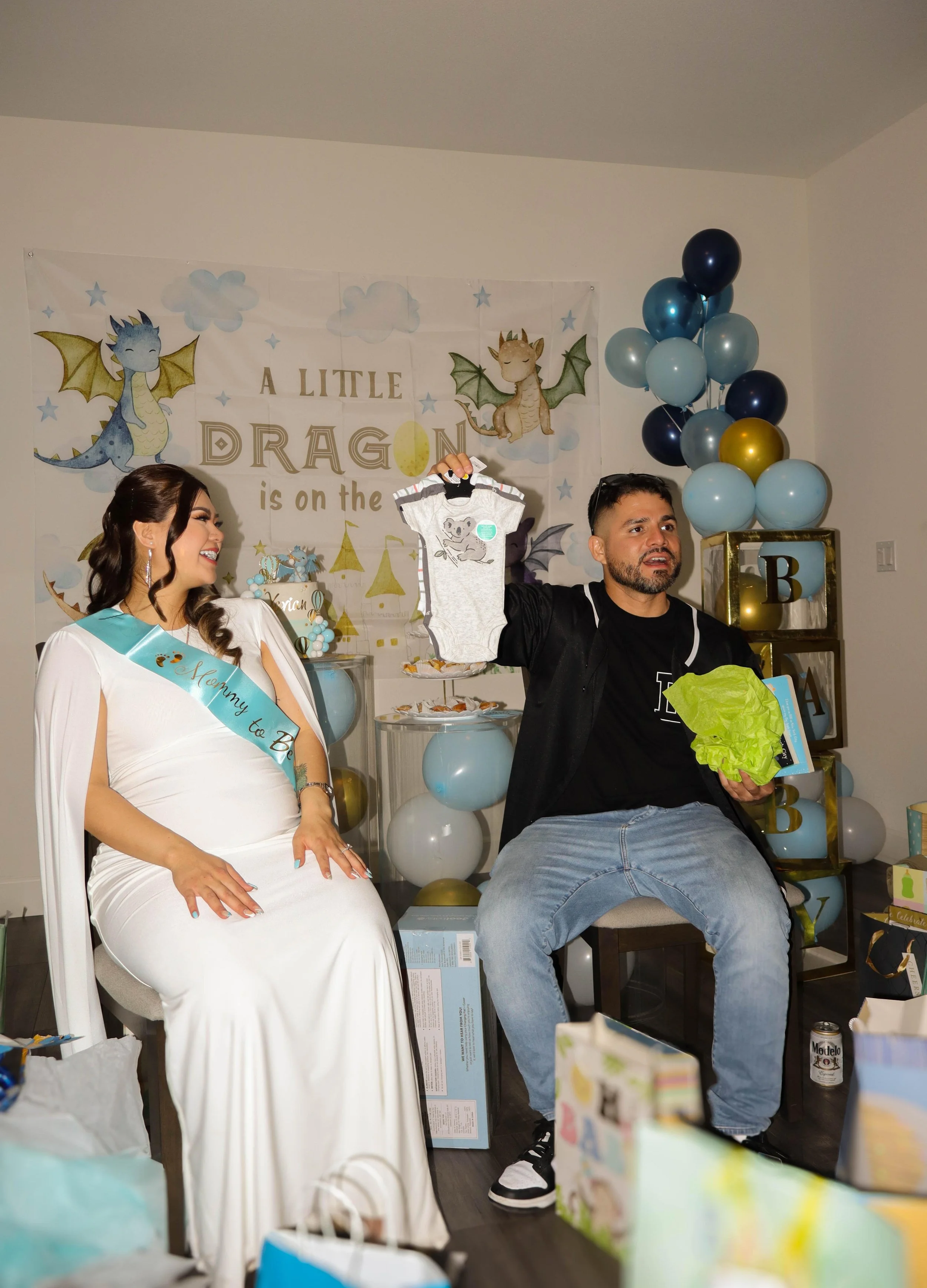 A woman dressed as a bride and a man are sitting indoors, celebrating a baby shower with a dragon theme. The man is holding up a baby onesie. Decorations include balloons, a dragon-themed backdrop, and gifts.