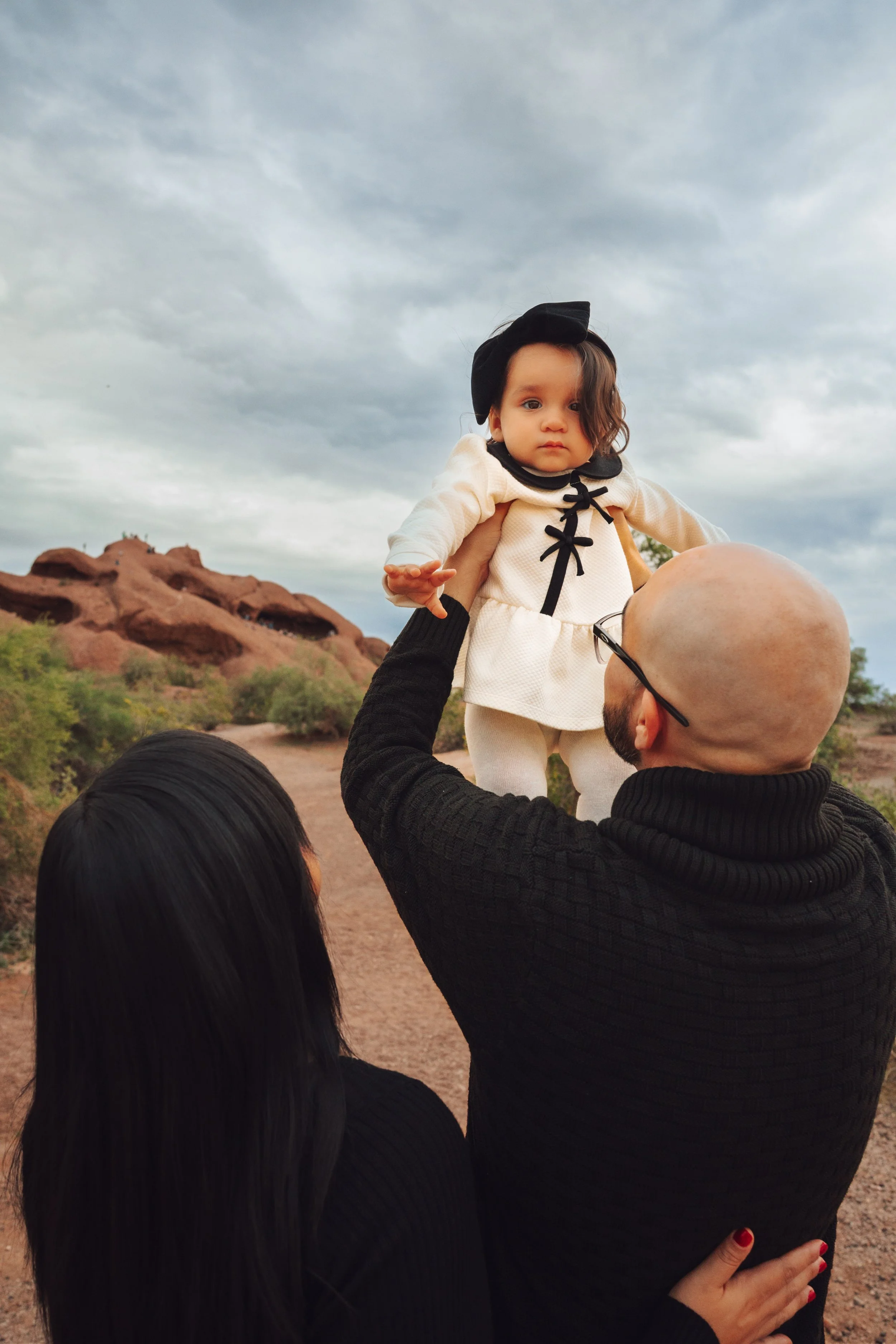 A man lifting a young girl in the air outdoors on a cloudy day with desert landscape and rock formations in the background.