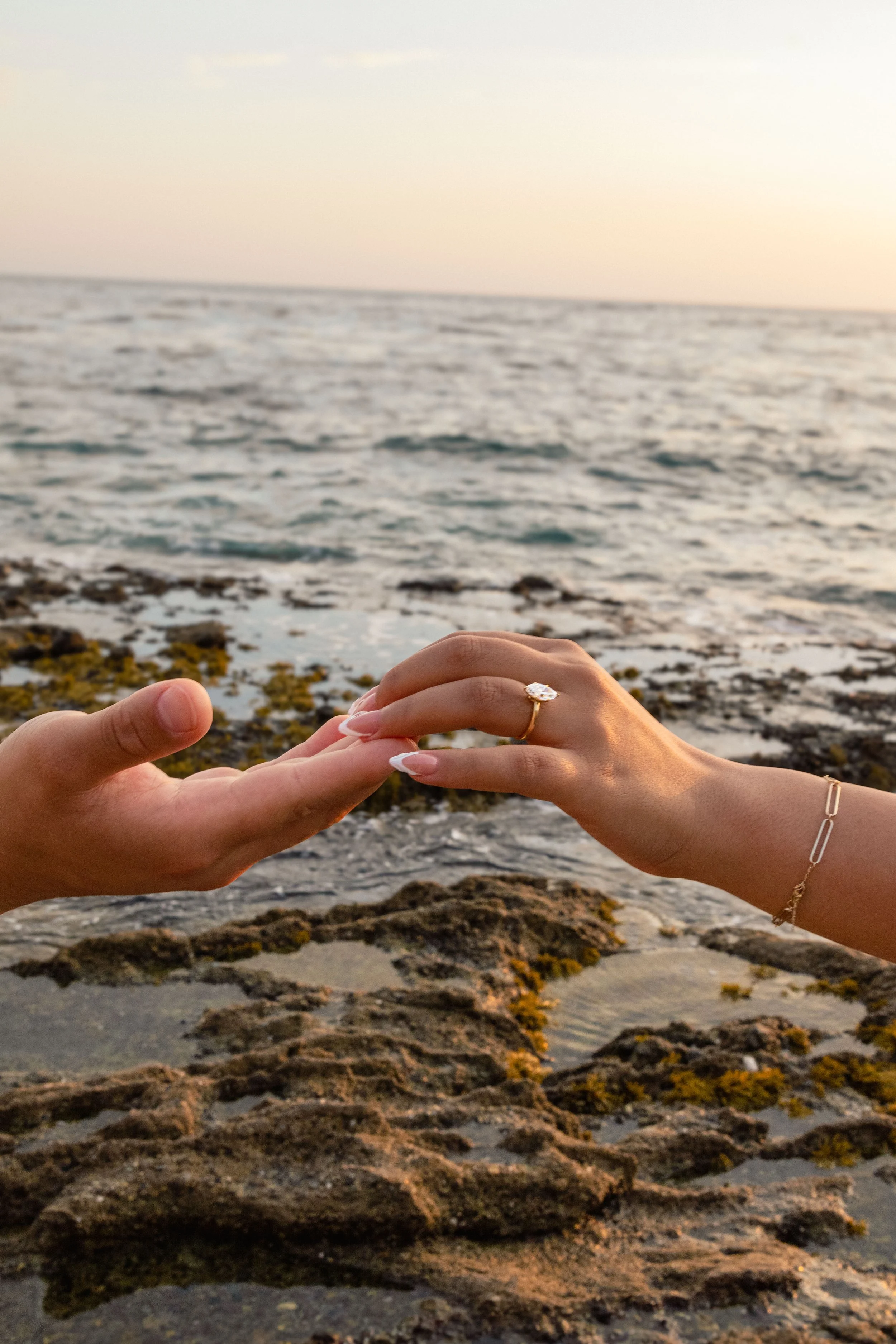 Two hands reaching towards each other, with a focus on a ring on one finger, against a backdrop of a rocky shoreline and ocean at sunset.